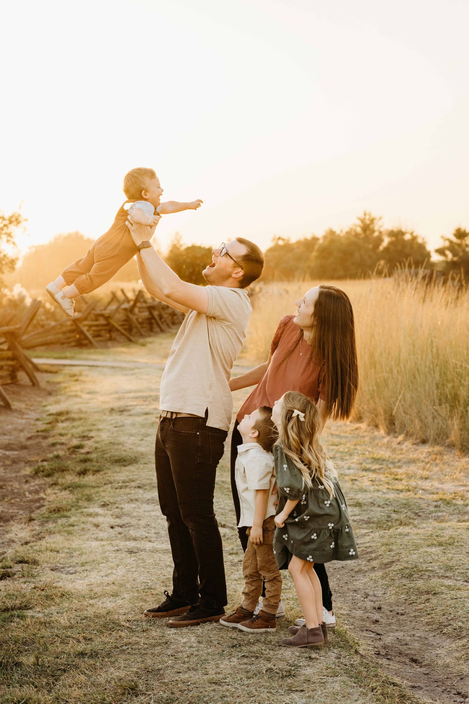A candid moment of a family of five with the dad holding his one year old in the air and everyone looking at him and smiling.