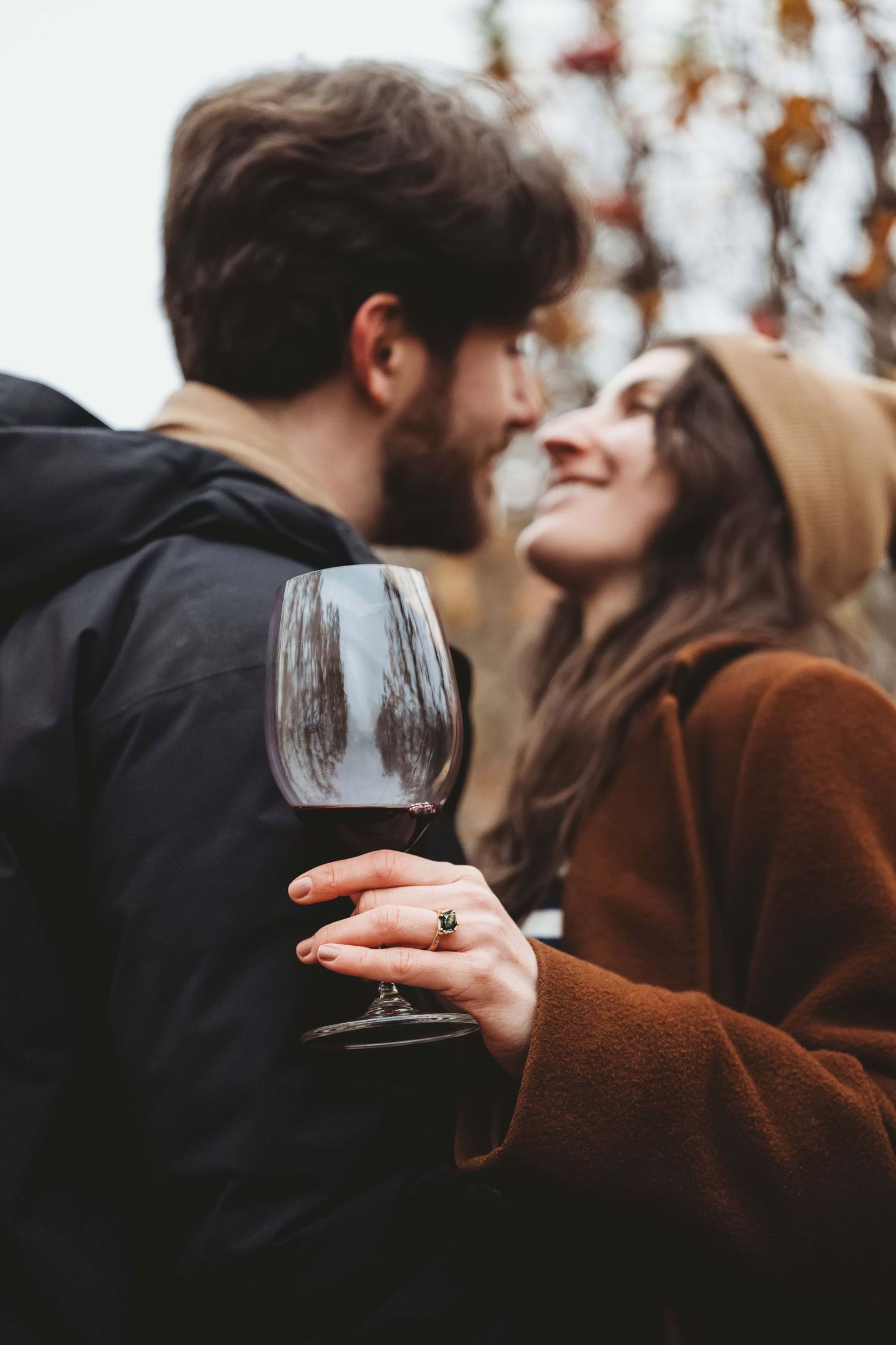 A couple looking at each other, smiling while standing in an apple orchard with a wine glass in hand.