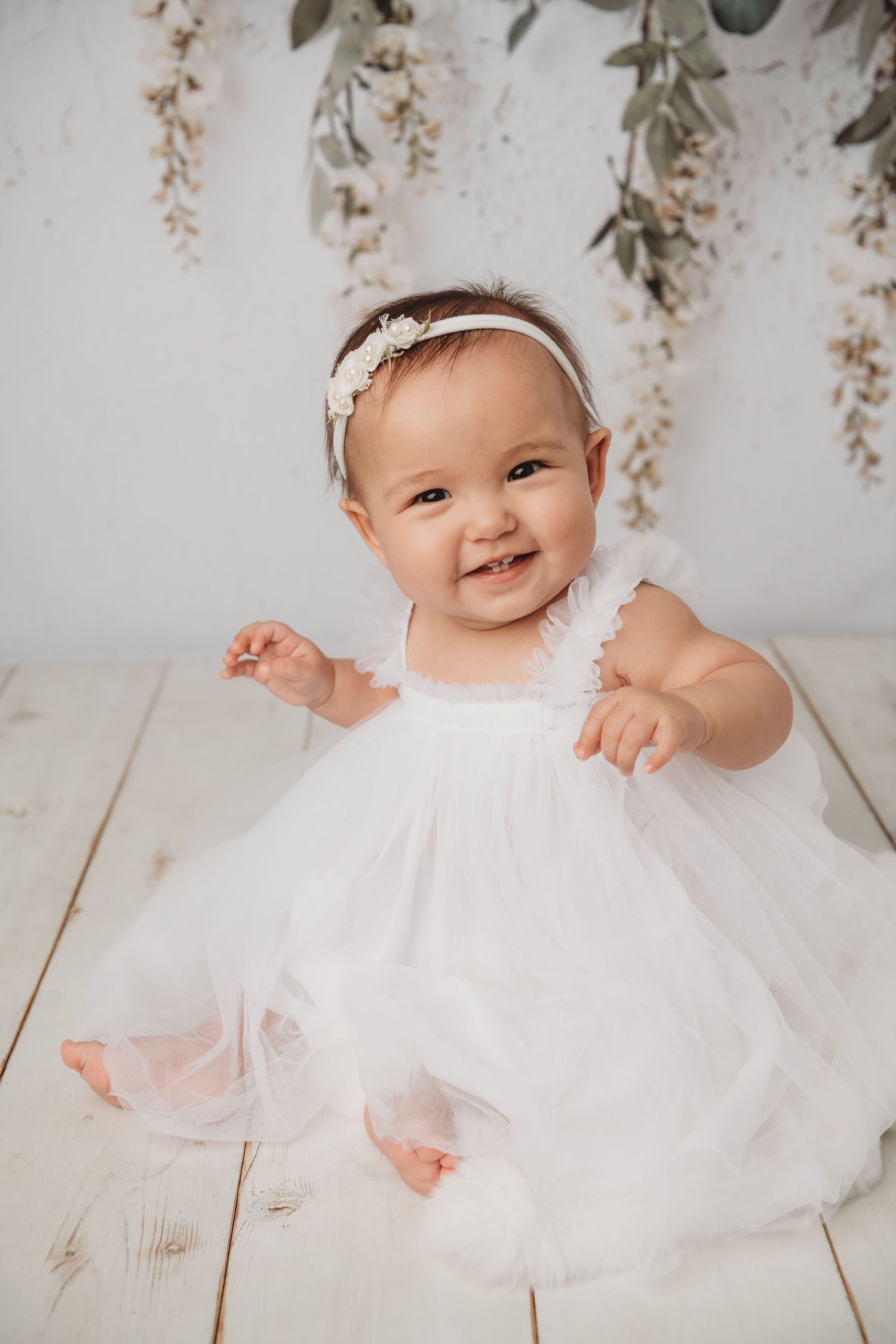 A baby sitting on a white wood floor wearing a white dress and smiling.