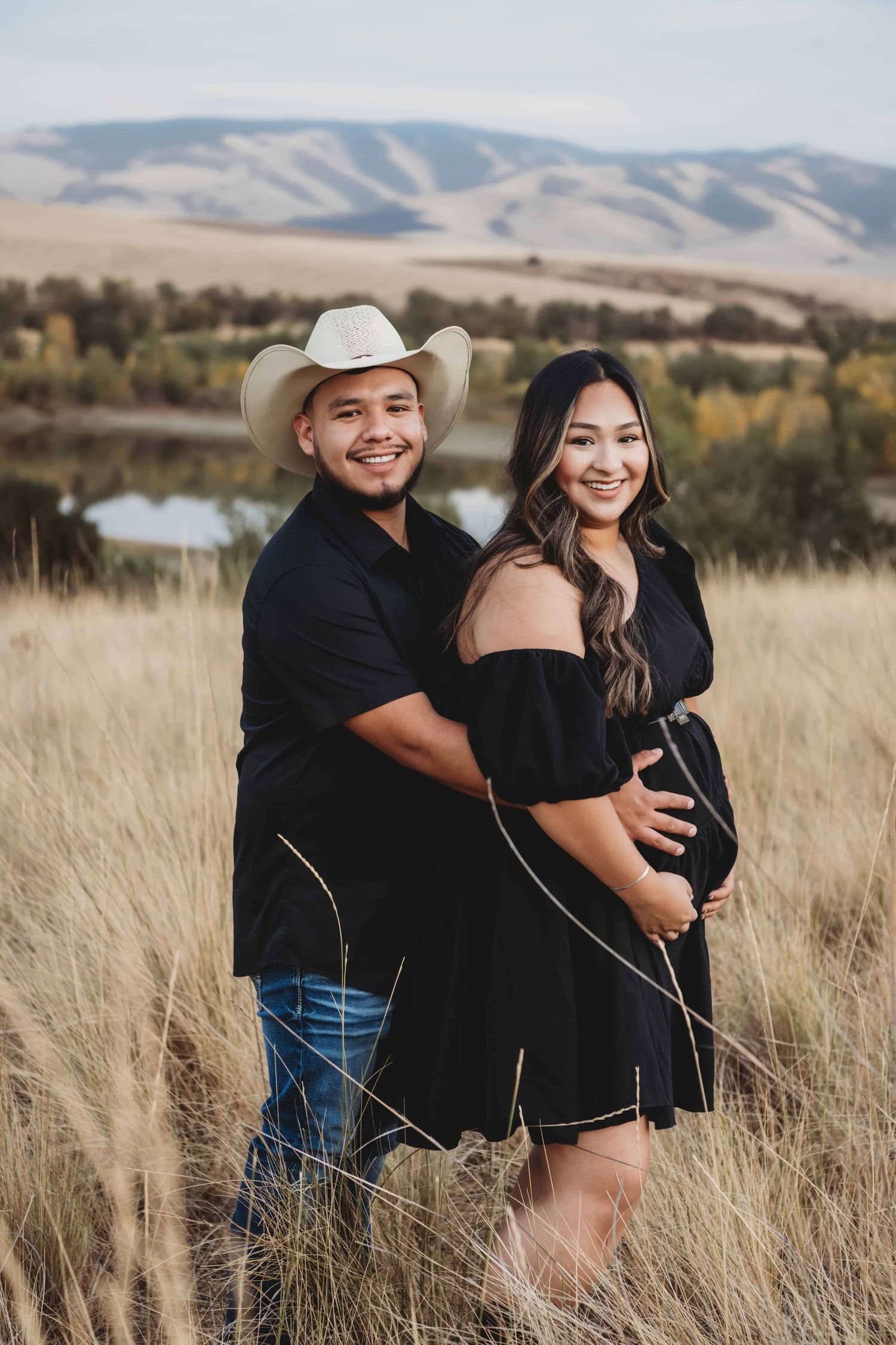 An expectant couple wearing black holding the pregnant belly with a lake and rolling hills in the background.