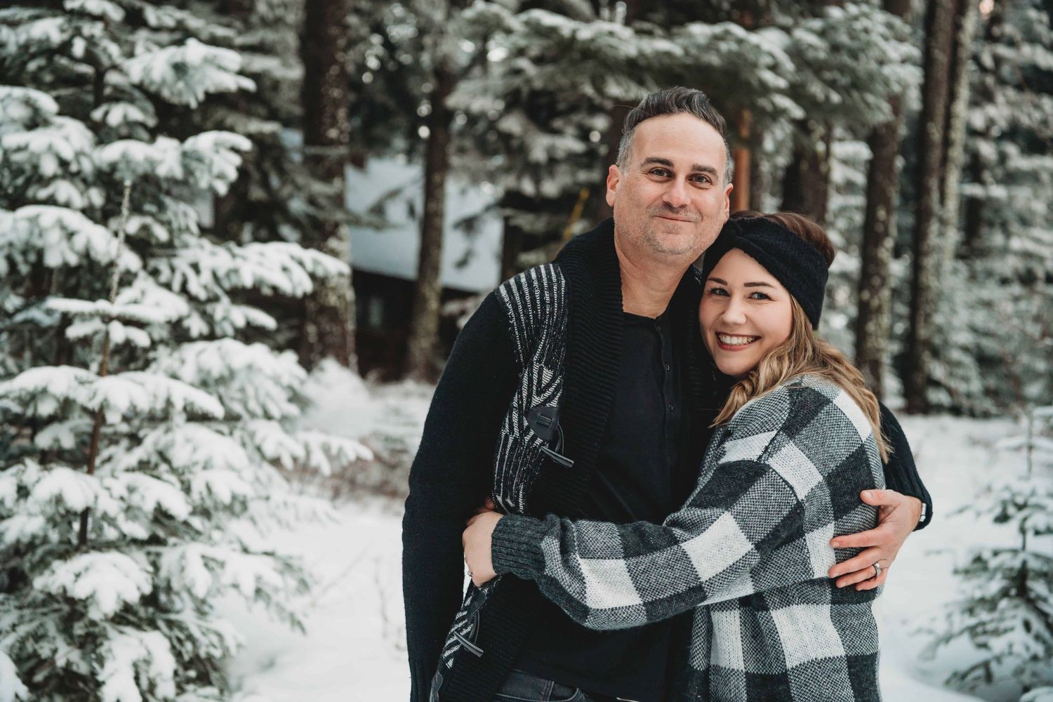A couple hugging each other while standing surrounded by tall trees and snow.