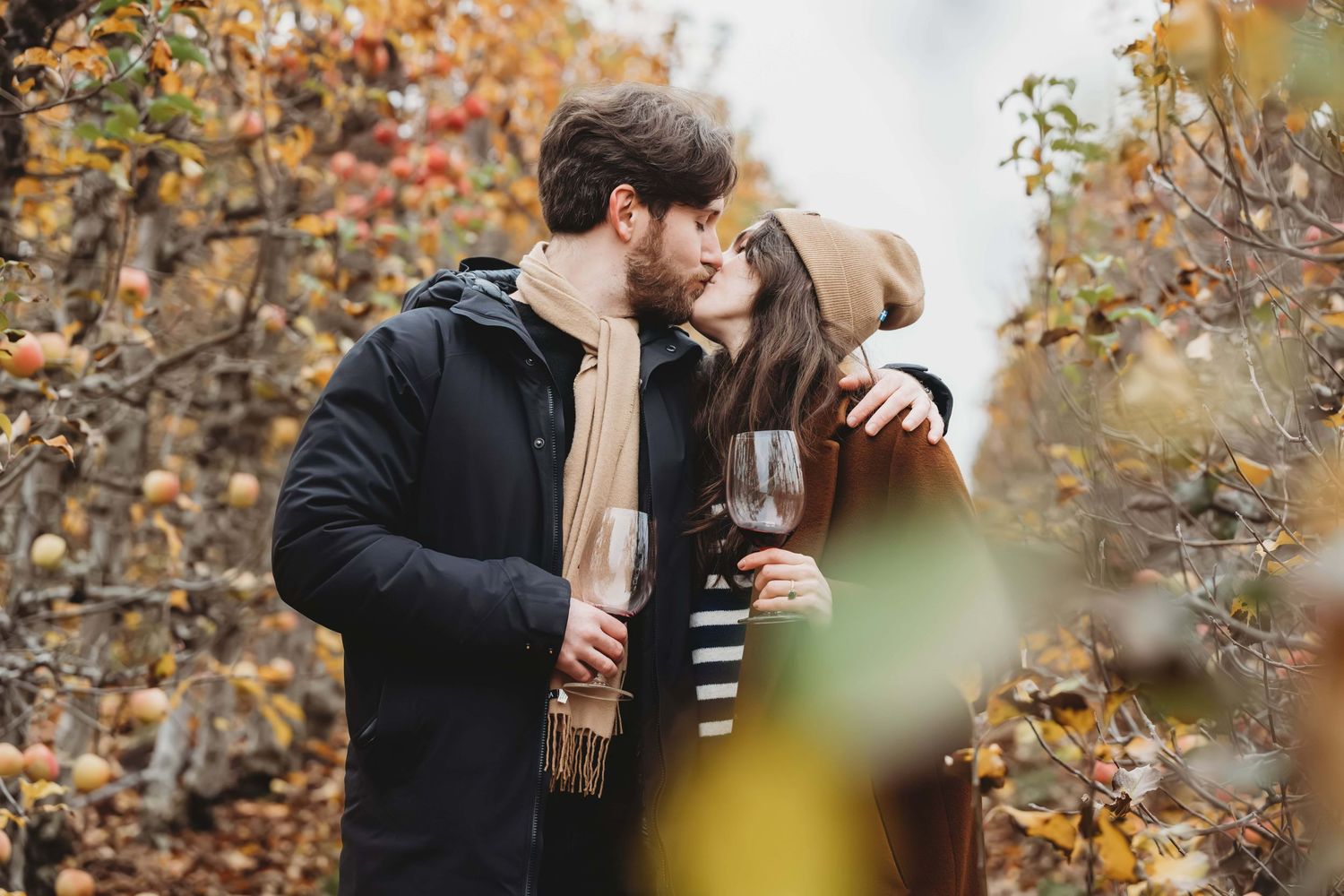 A couple kissing in an orchard with wine glasses in hand on a winter day.