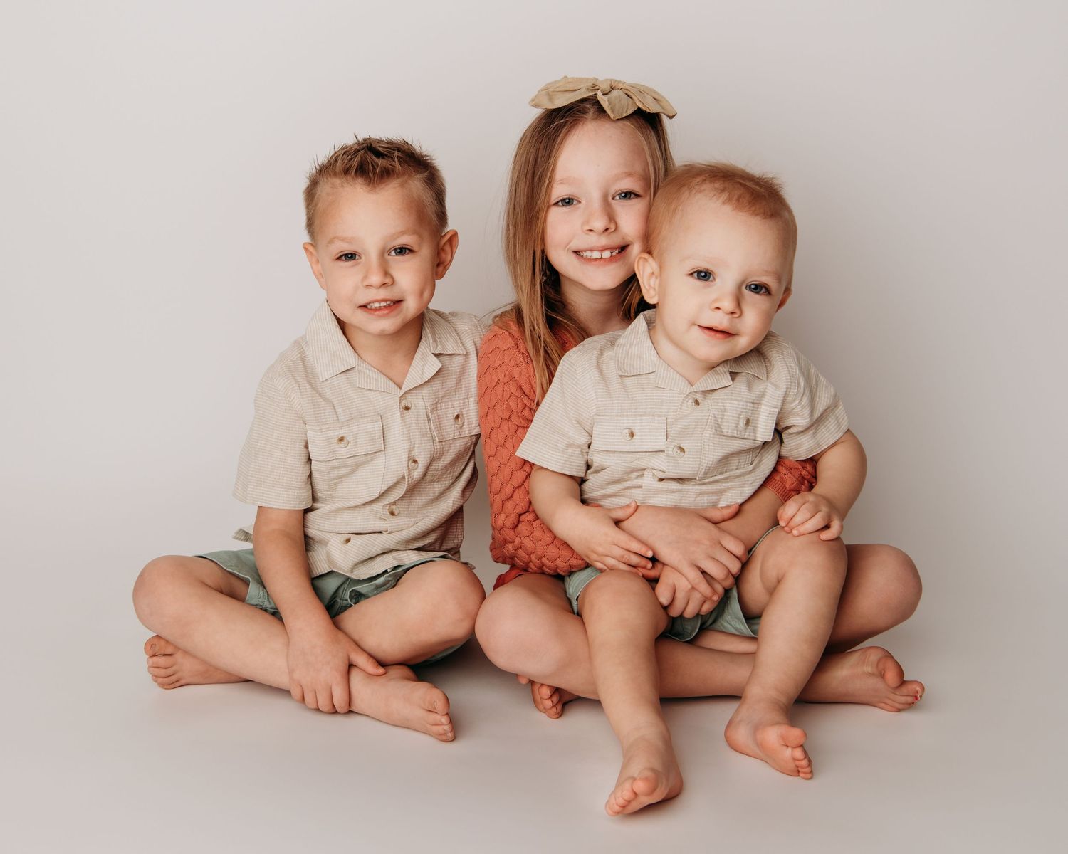 A Walla Walla photographer captures three siblings sitting on a white backdrop smiling for the camera.