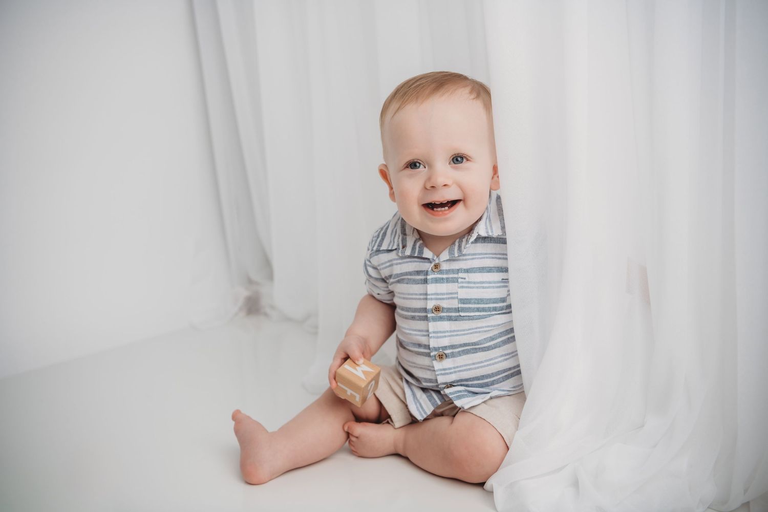 A baby sitting between sheer curtains laughing and holding a block in his hand.