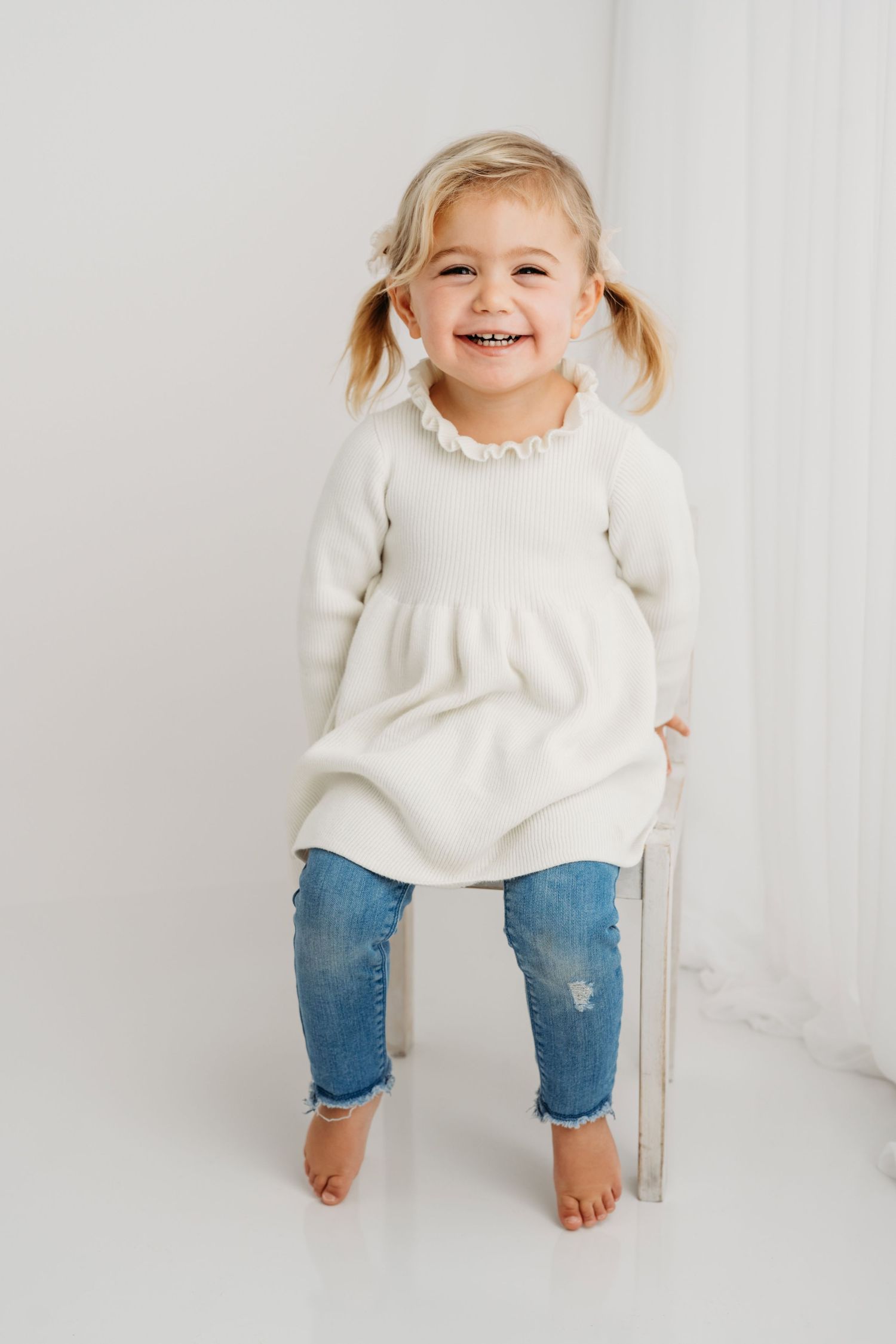 A classic portrait of a child sitting on a white chair wearing a white shirt and jeans.