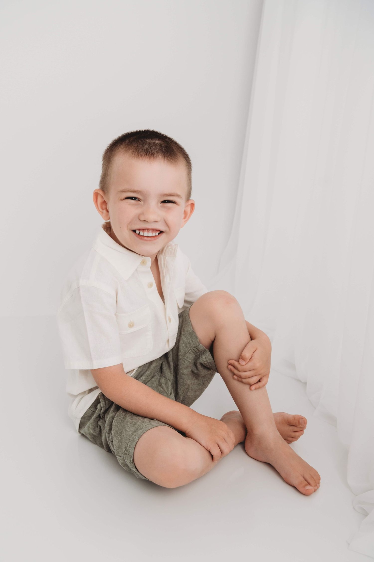 A timeless portrait of a boy sitting on a white backdrop with his knee up and smiling.