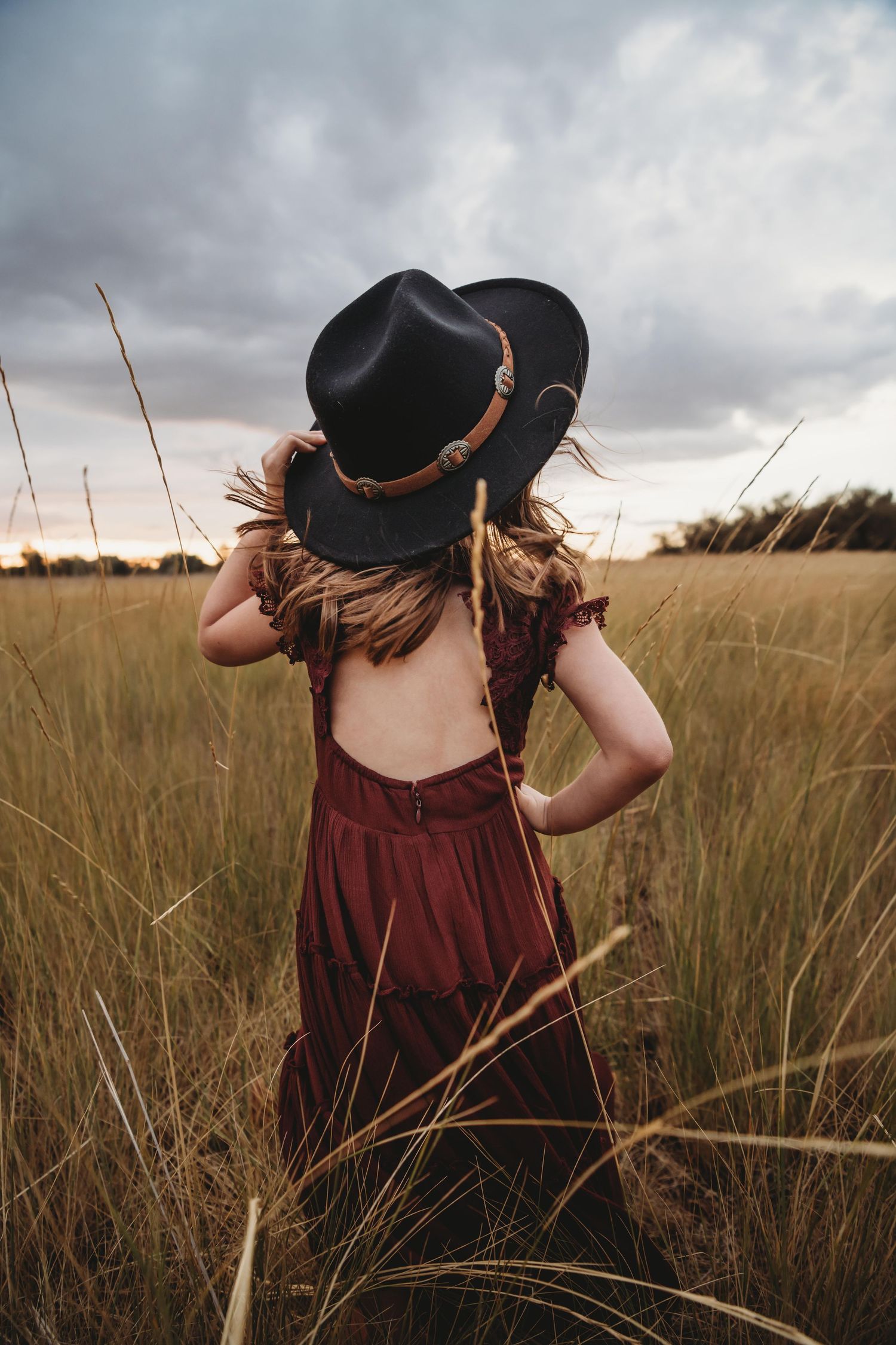 A candid portrait of a girl standing in a field with a hat on during sunset on a windy, stormy day in Walla Walla.