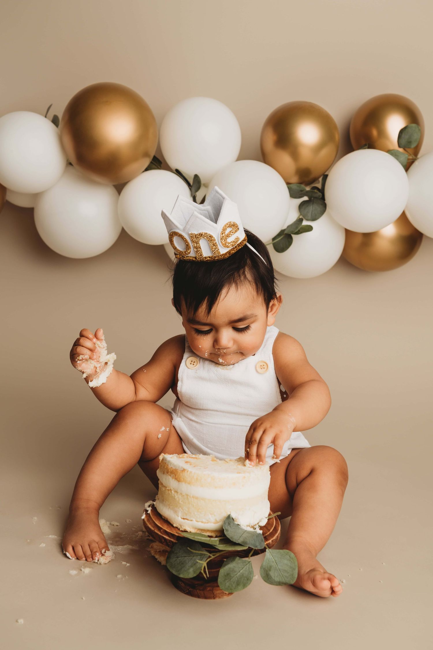 Studio portrait in Walla Walla of a one year old baby eating a white cake on a tan backdrop with balloons.