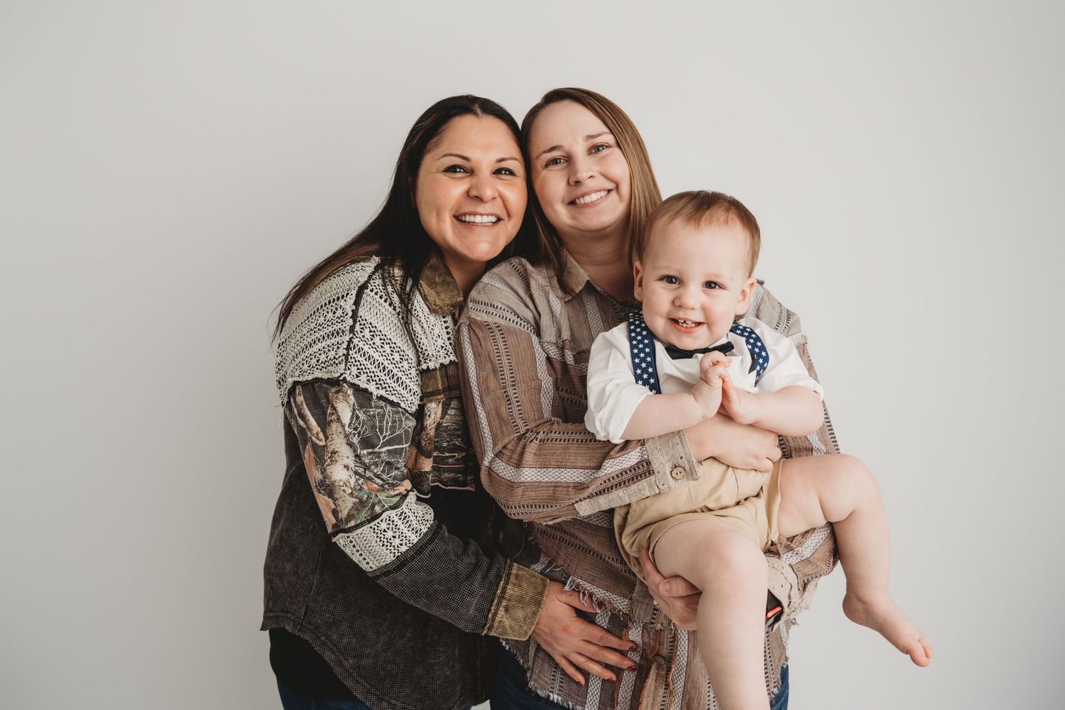 Two moms hold their one year old boy in a timeless studio family portrait.