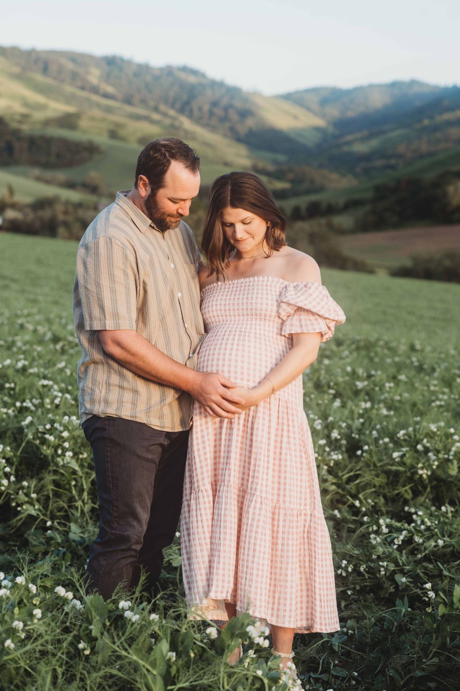An expecting couple standing in a pea field with green hills in the background.
