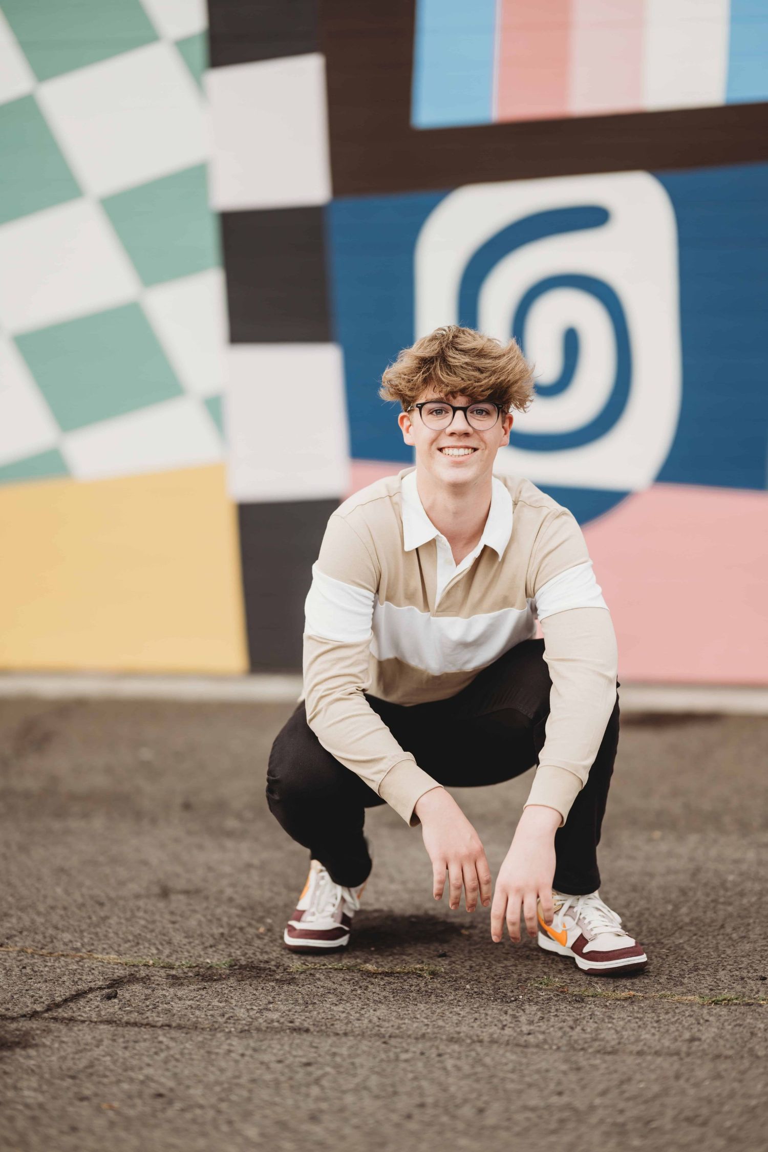 A senior portrait of a young man squatting in front of some artwork on a wall in downtown Walla Walla.