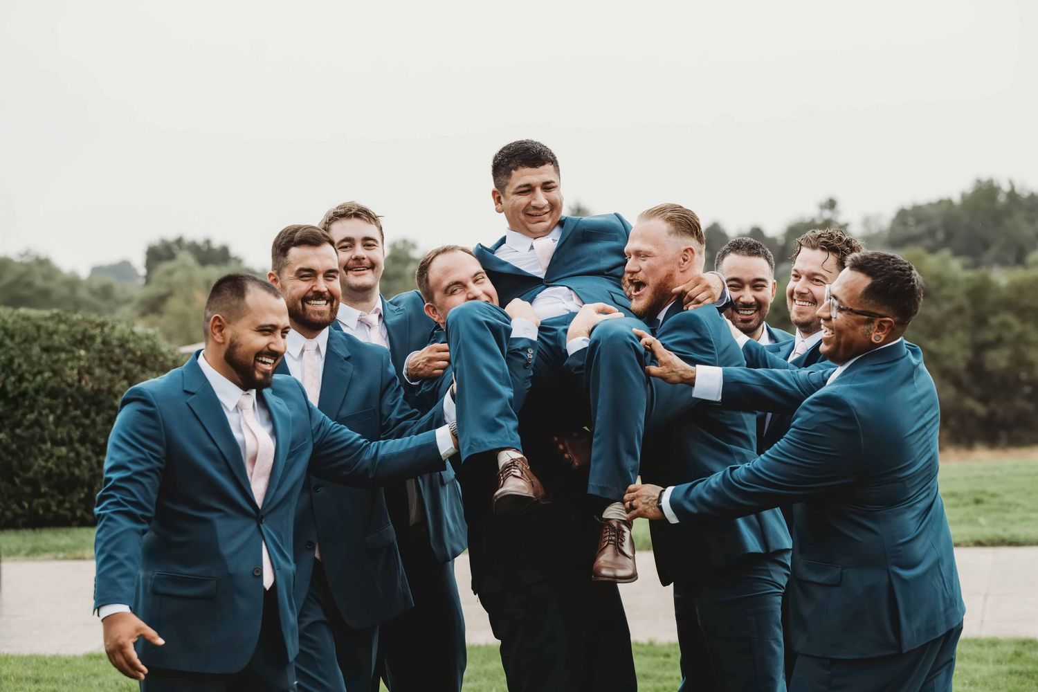 A group of groomsmen picking up the groom, all dressed in navy suits.