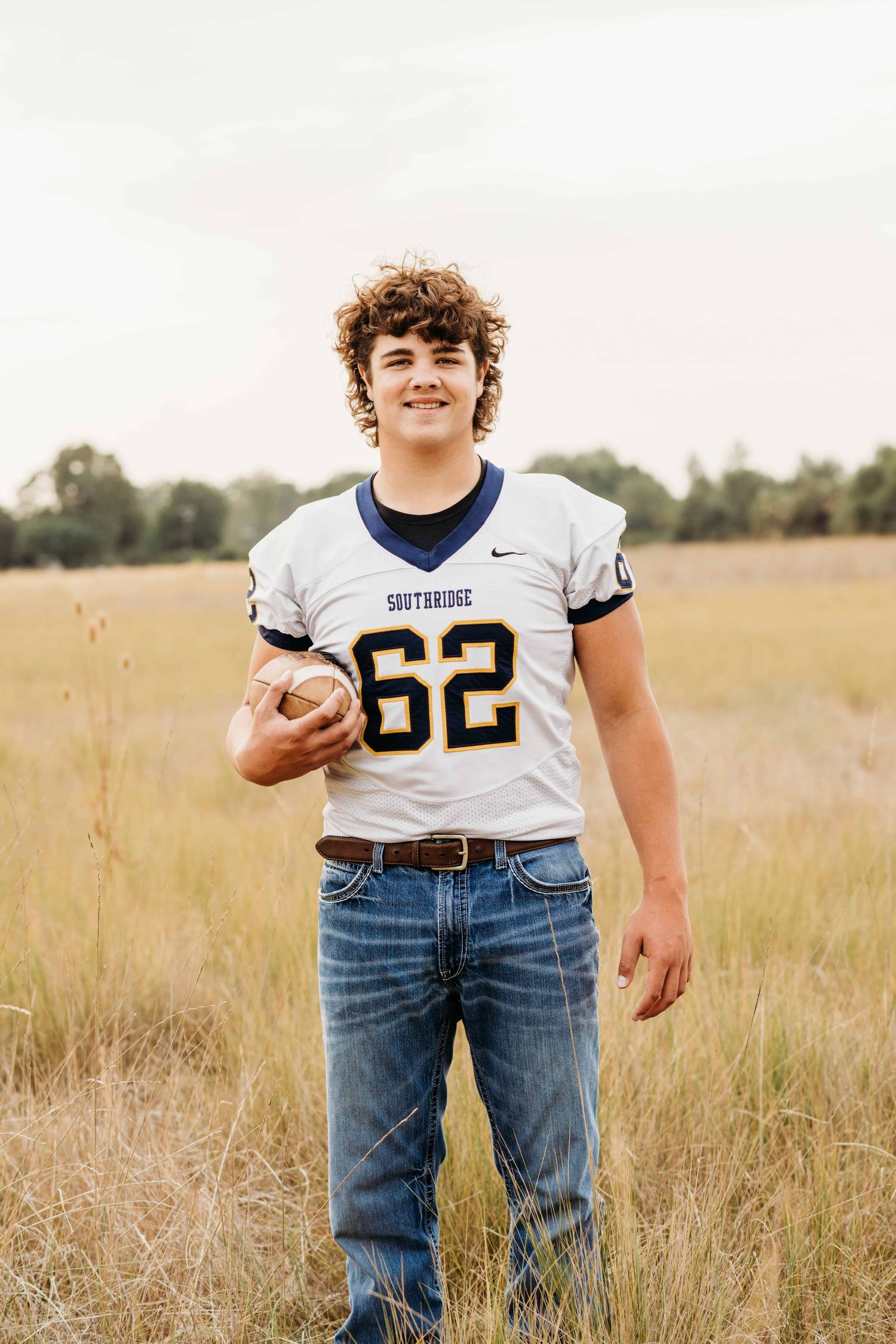 A high school senior in a football uniform, holding a football while he smiles for the camera during his senior photo session in Walla Walla