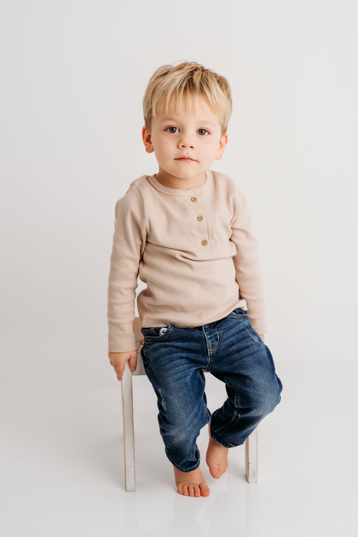 A boy sits on a white wooden chair with a serious expression on his face.