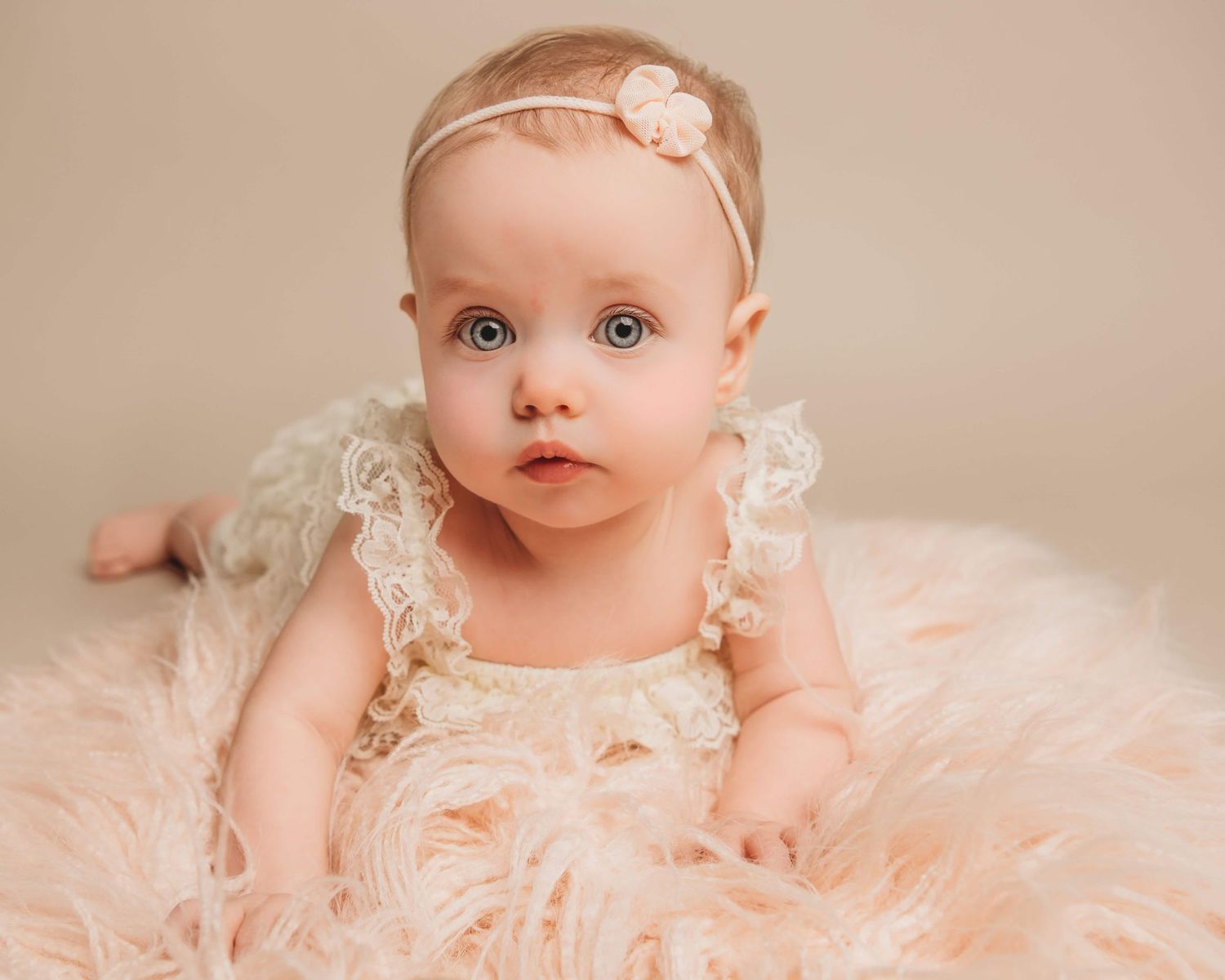 A baby girl with big blue eyes laying on a soft pink rug with a serious expression.