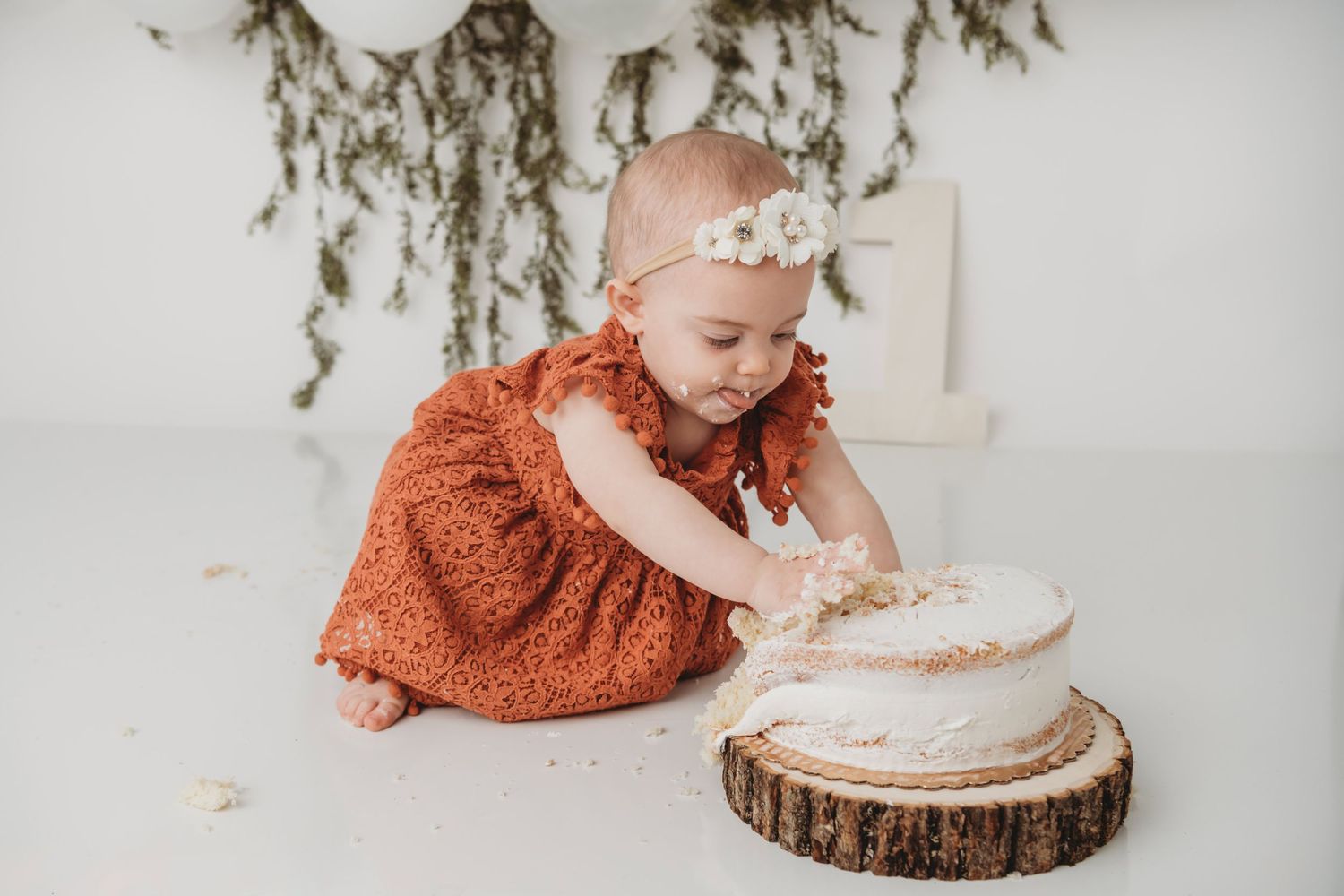 A one year old girl reaches for her white cake on a wooden cake stand.