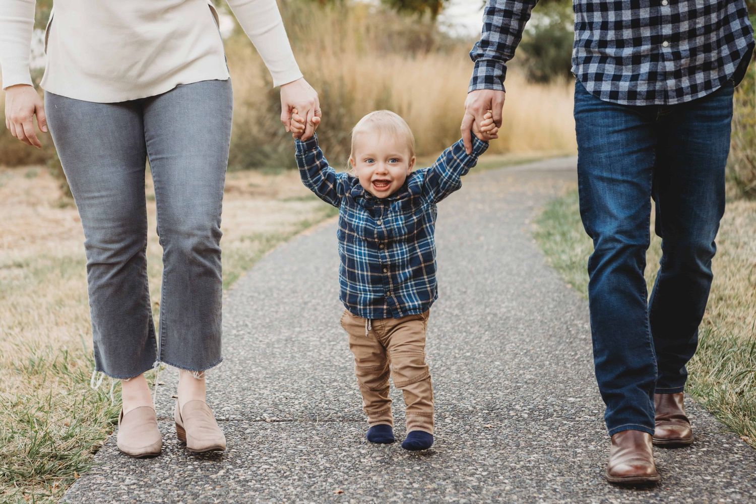 A baby walks on a path holding his parents hands in Walla Walla.