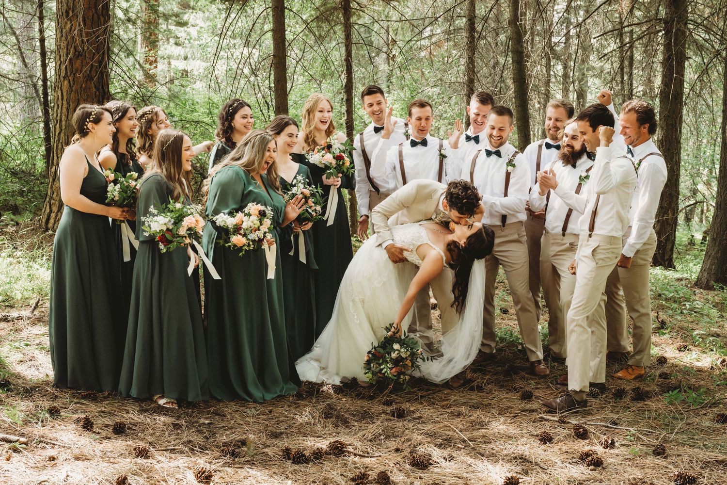 A wedding party cheering on the bride and groom as they kiss in a wooded area.