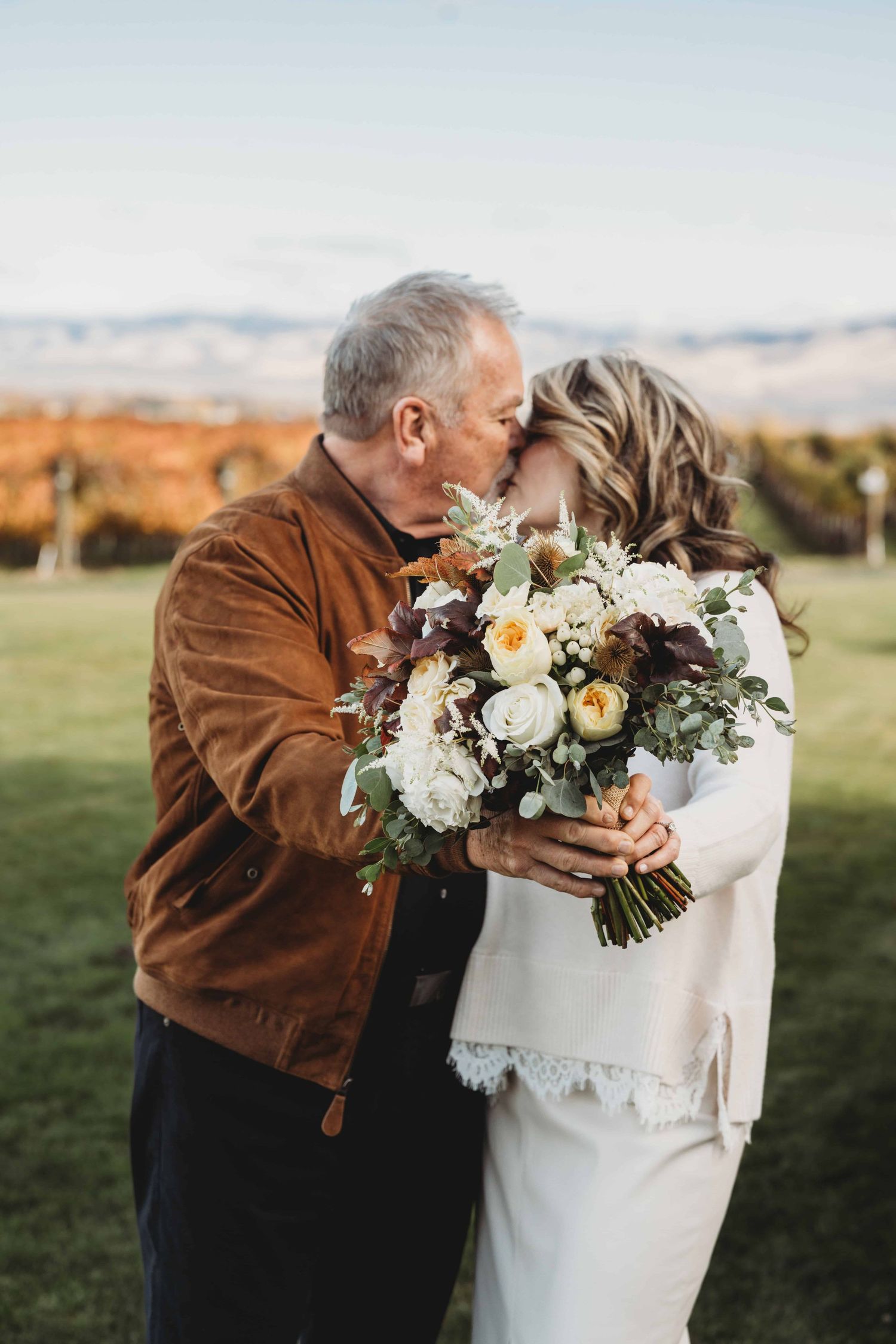A bride and groom kissing and holding out their fall colored bouquet in front of them.