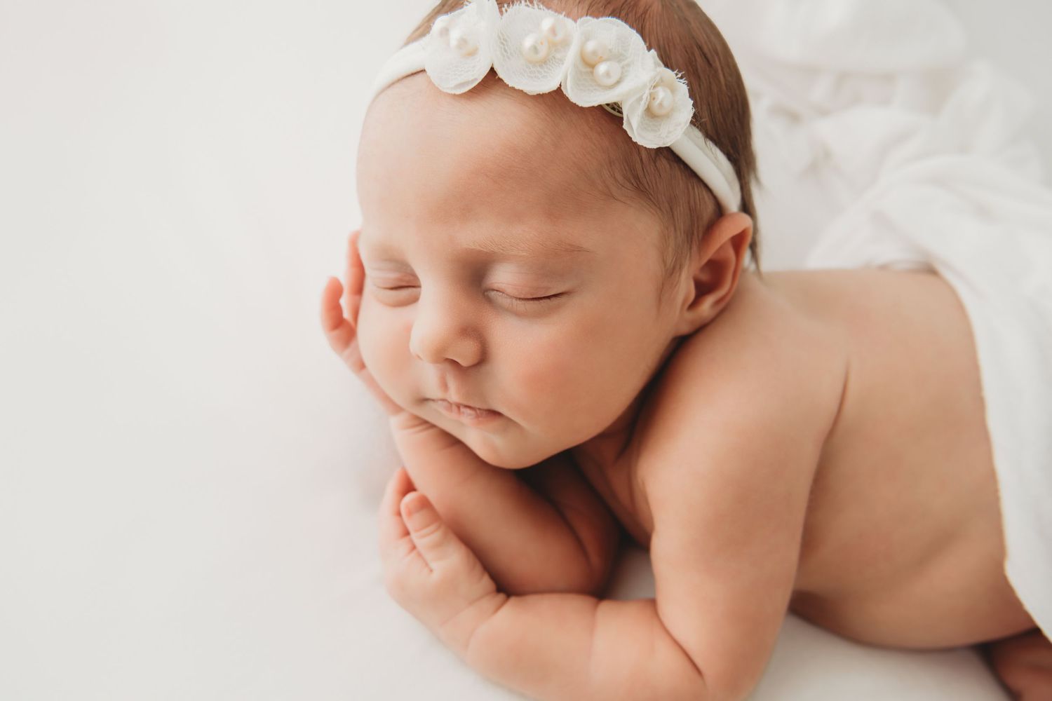 A newborn baby girl sleeping with her arms under her face wearing a white pearl headband on a white blanket.