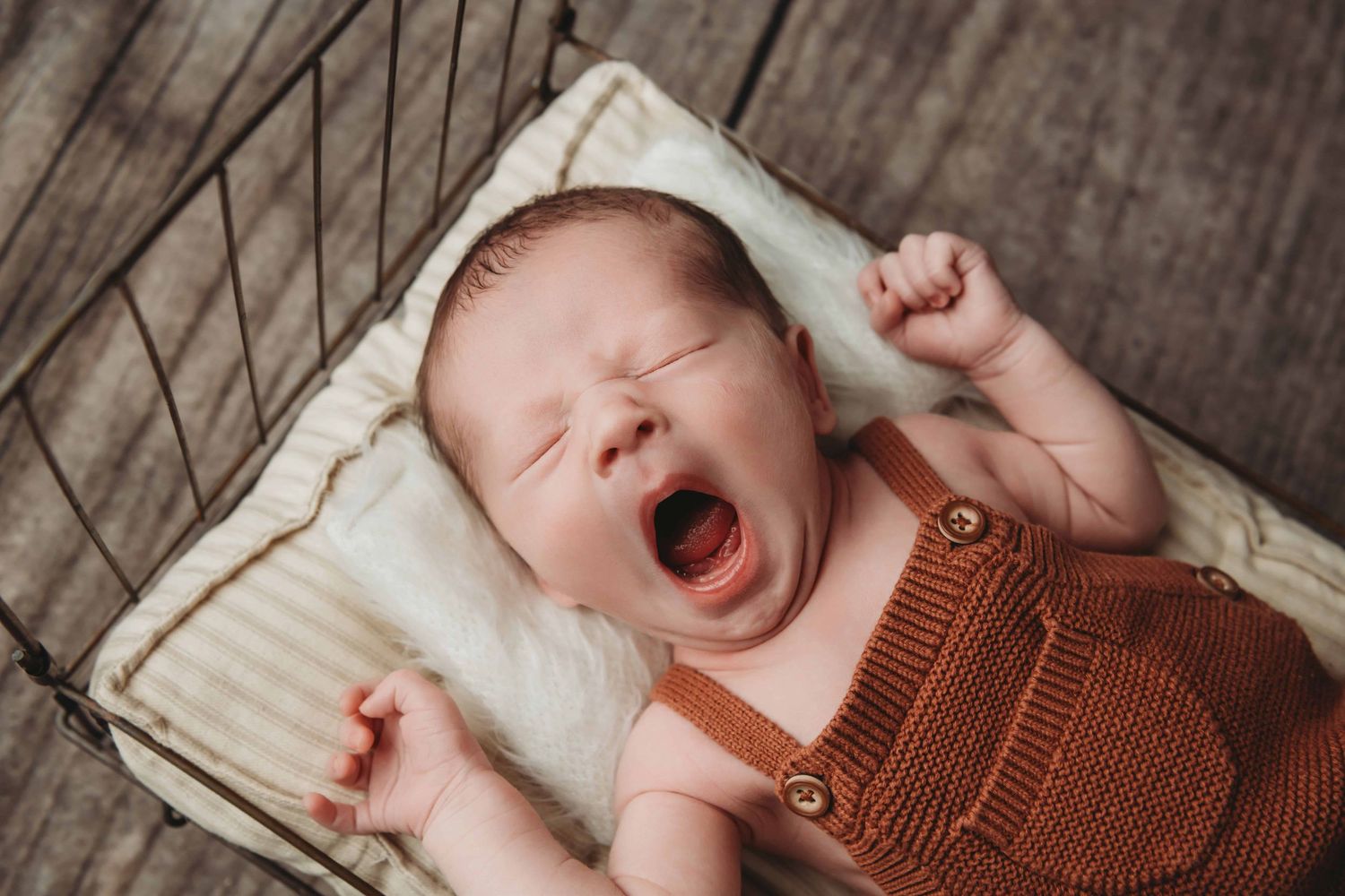 A baby boy yawning and stretching while laying on a metal bed with a striped mattress.