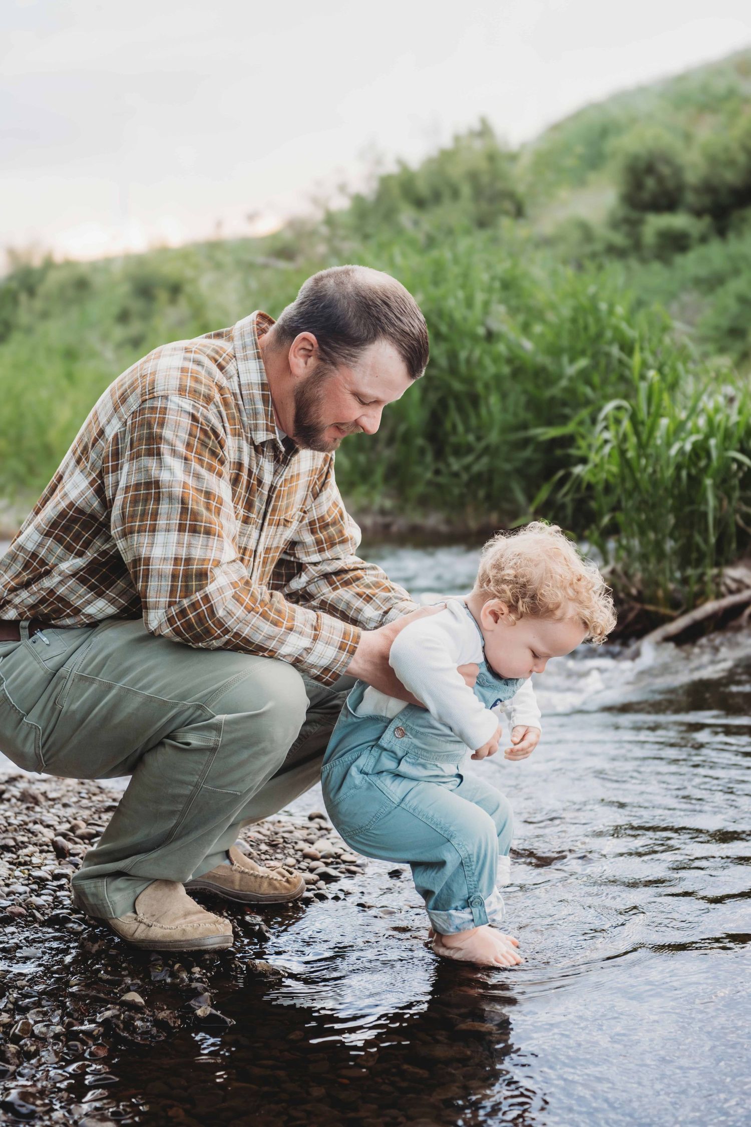 A father holding his baby and letting him dip his feet in a shallow creek.