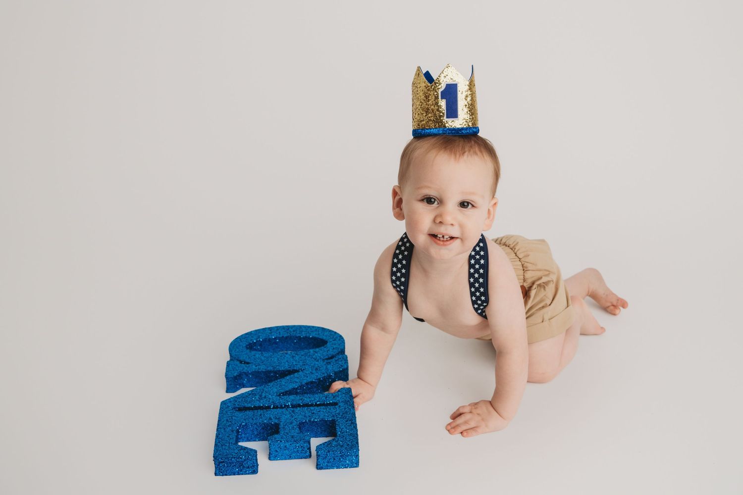 A one year old crawling on a white backdrop with a one crown on his head.