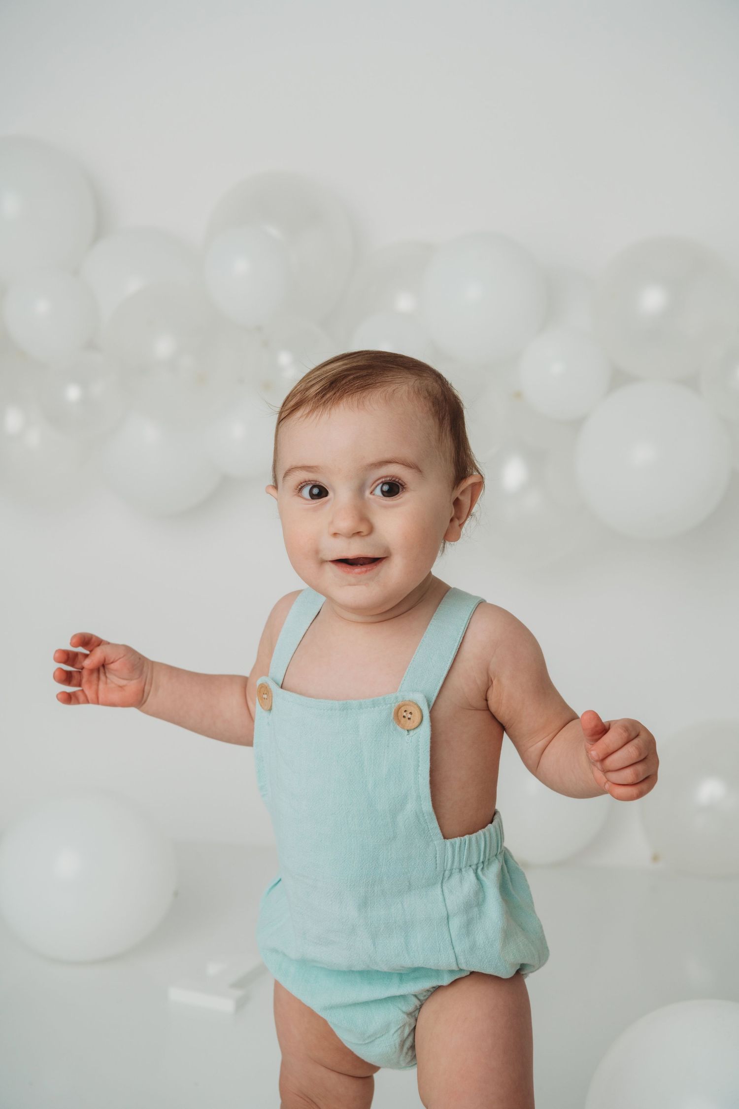 A one year old boy wearing a baby blue romper standing on a white backdrop with clear and white balloons.