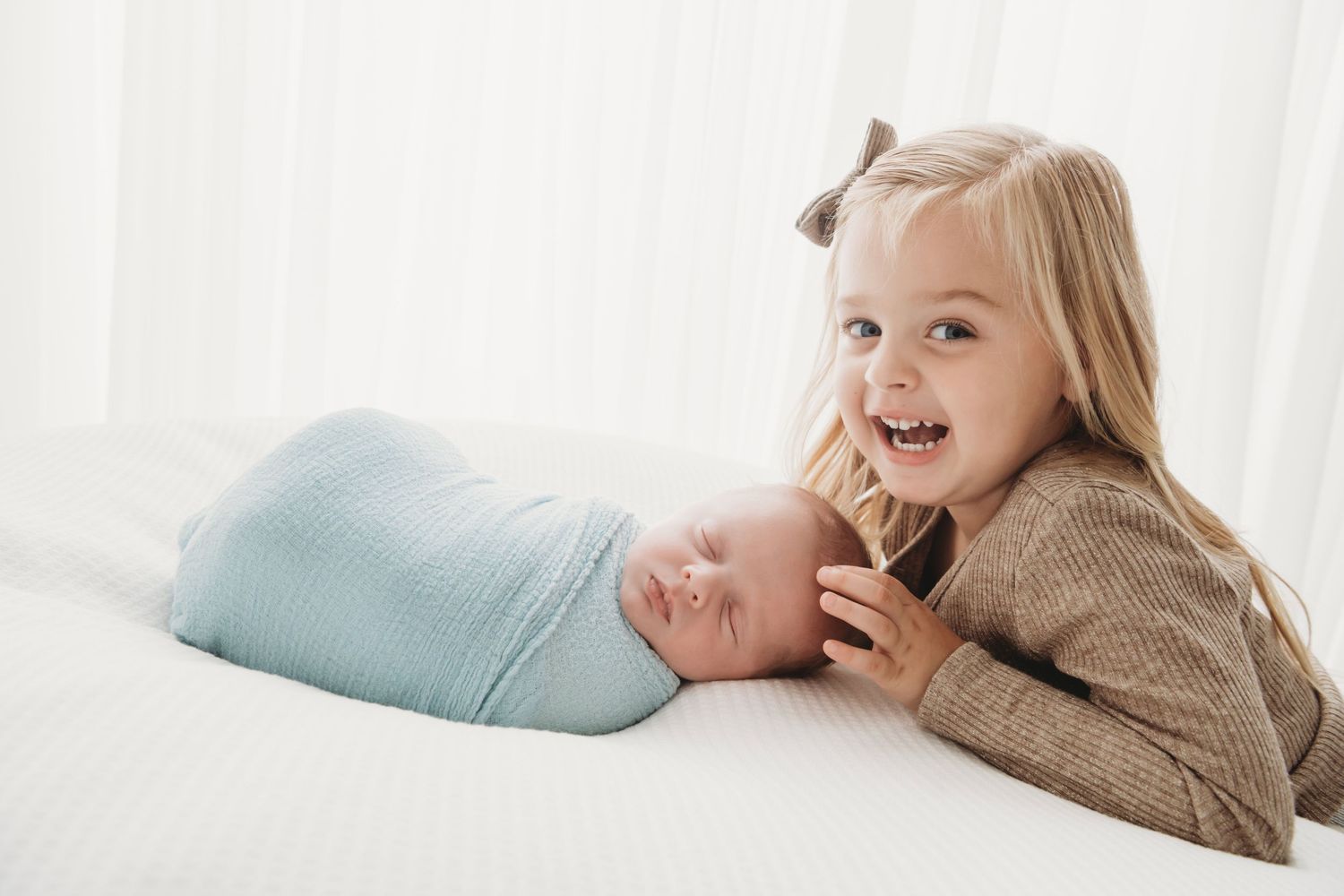 A backlit photo of a girl leaning in and touching her newborn brother's head.
