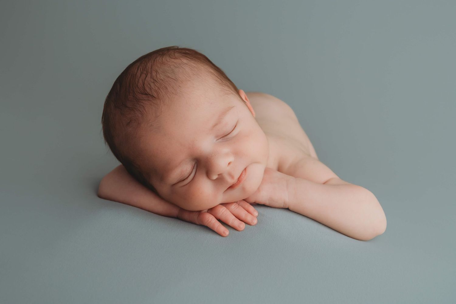 Studio newborn portrait of a baby sleeping on a blue blanket with his hands by his face.