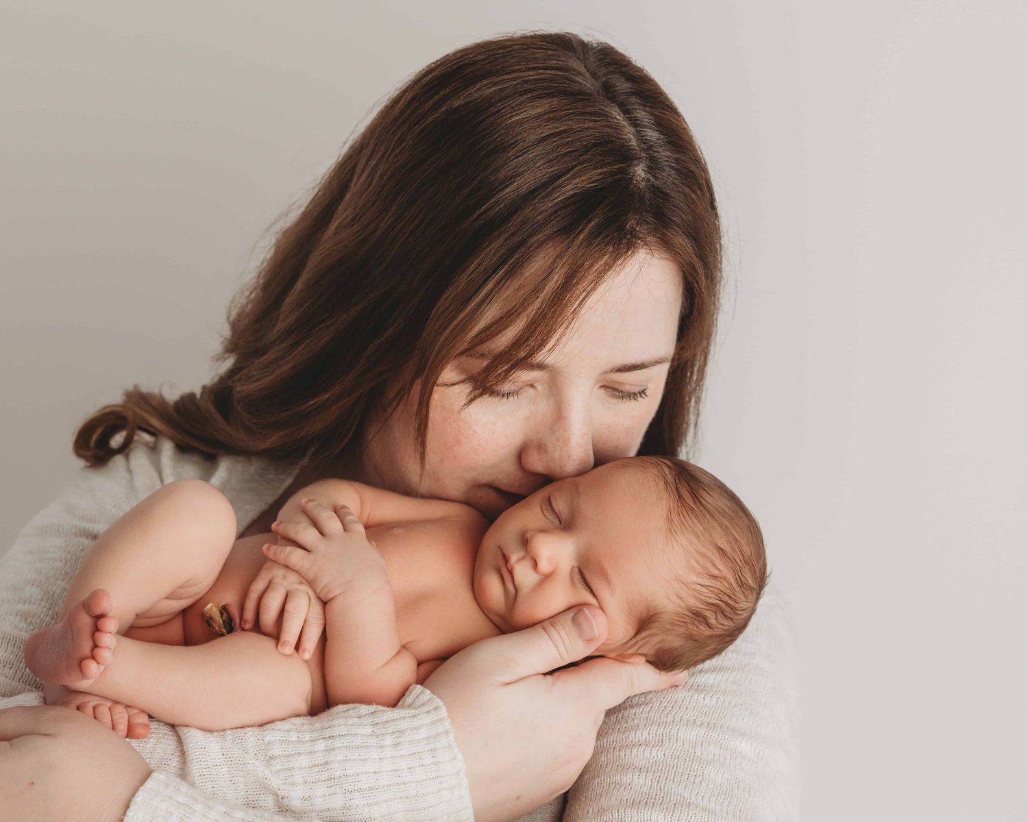A mother holding and kissing her newborn baby on a neutral background.
