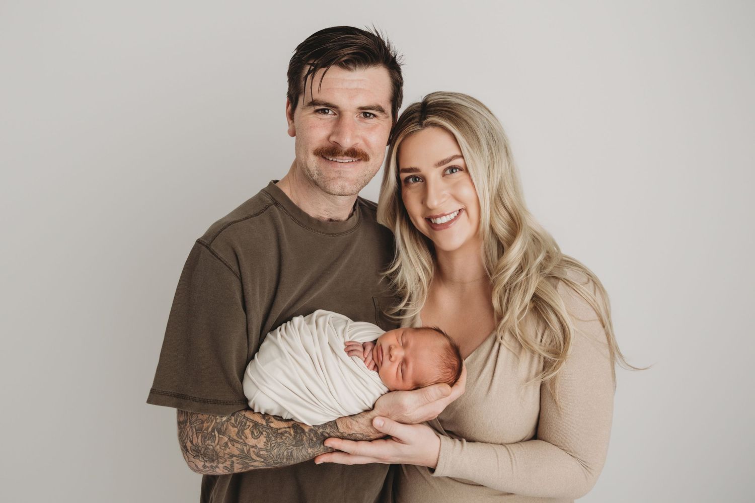New parents holding their sleeping newborn wrapped in white on a neutral backdrop, smiling for the camera.