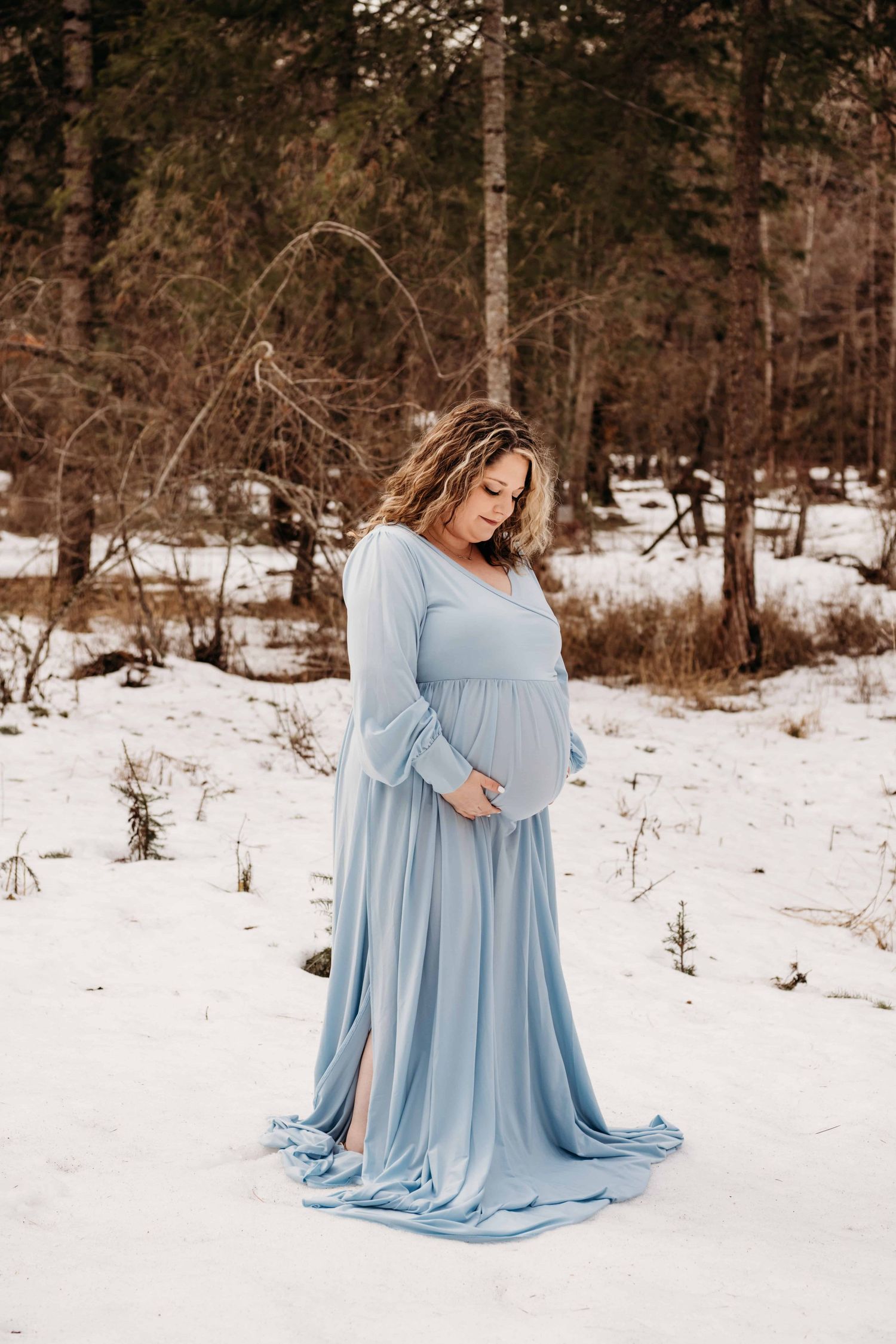 A pregnant woman standing in snow, wearing a baby blue dress looks down at her belly with a soft smile.