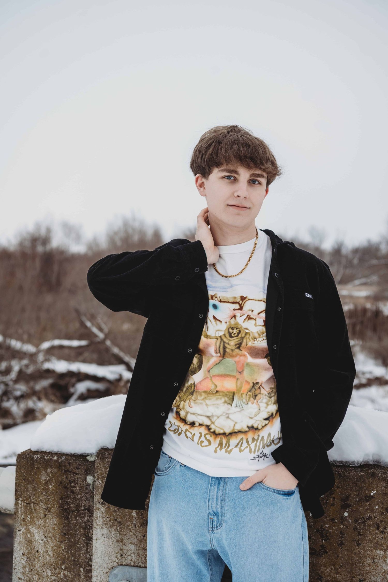 A senior portrait of a young man standing and looking at the camera with snow in the background.