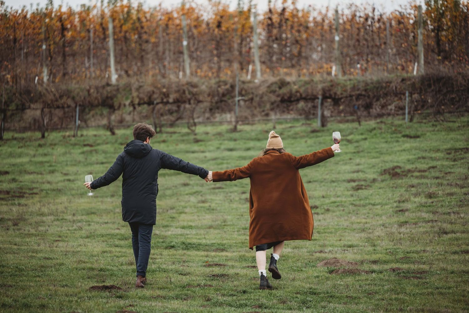 A couple walking through a grassy field with wine glasses in hand.