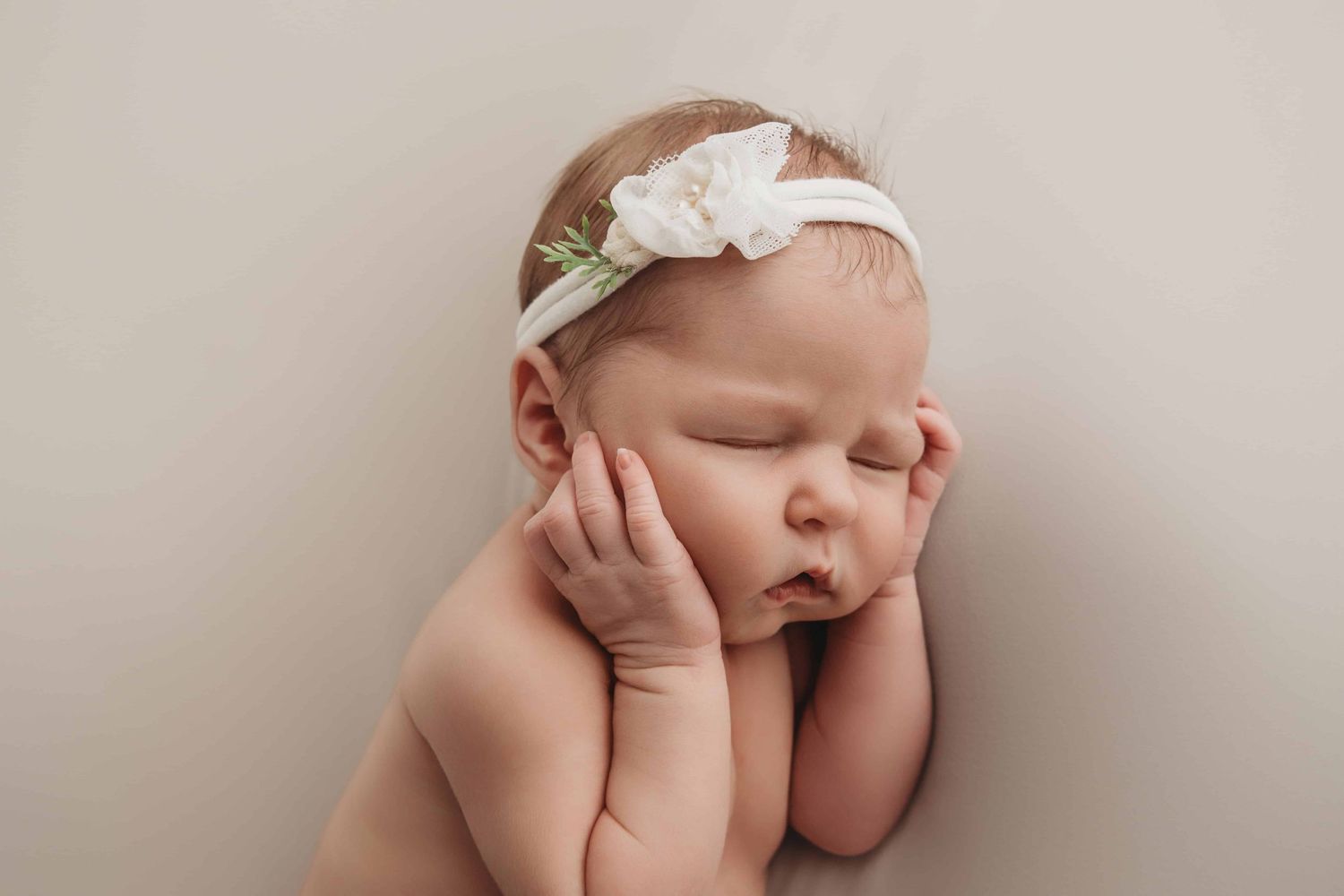 A newborn with chubby cheeks sleeping on a neutral blanket wearing a white headband.