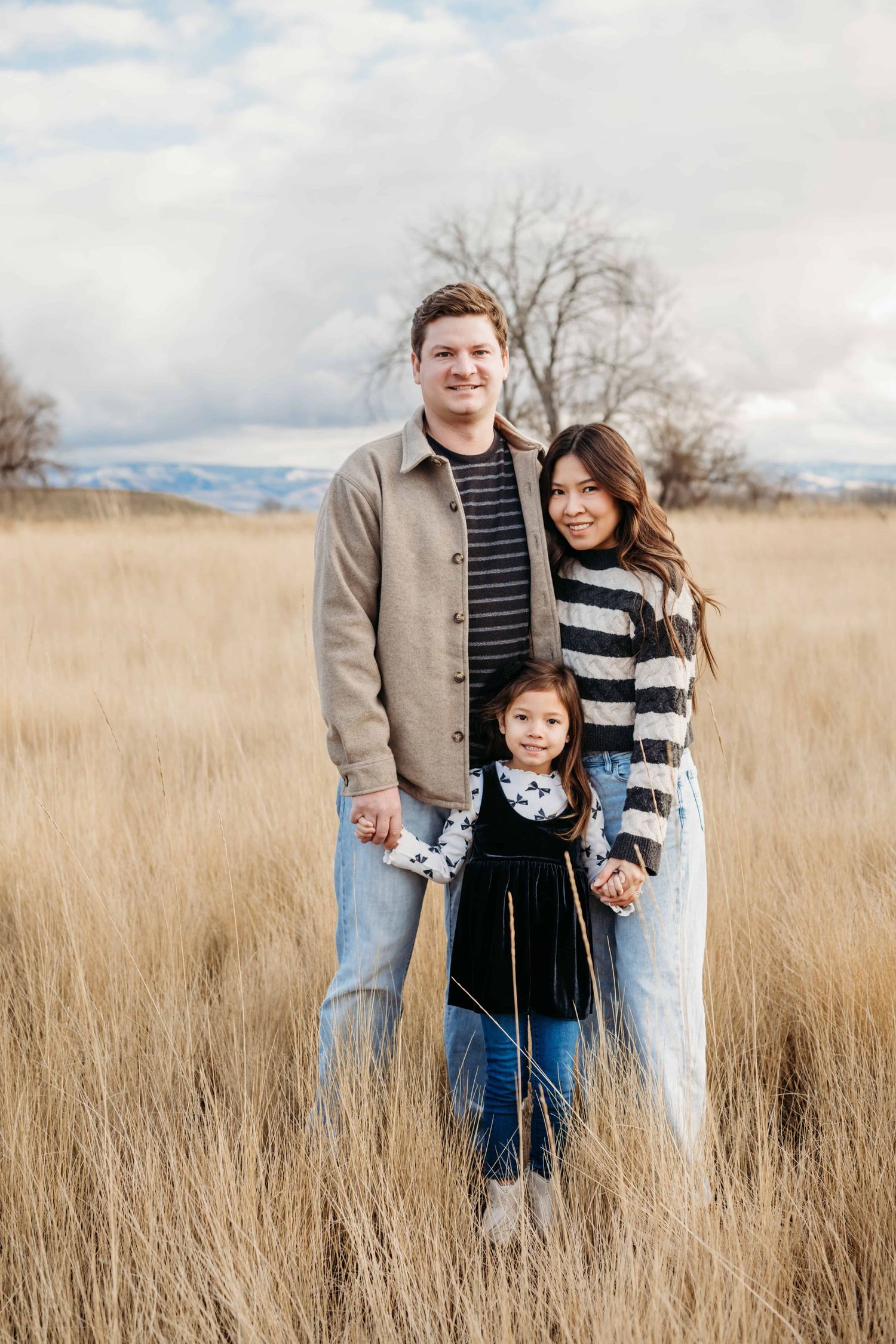 A portrait of a family standing in a field on a cloudy day wearing neutral colors.