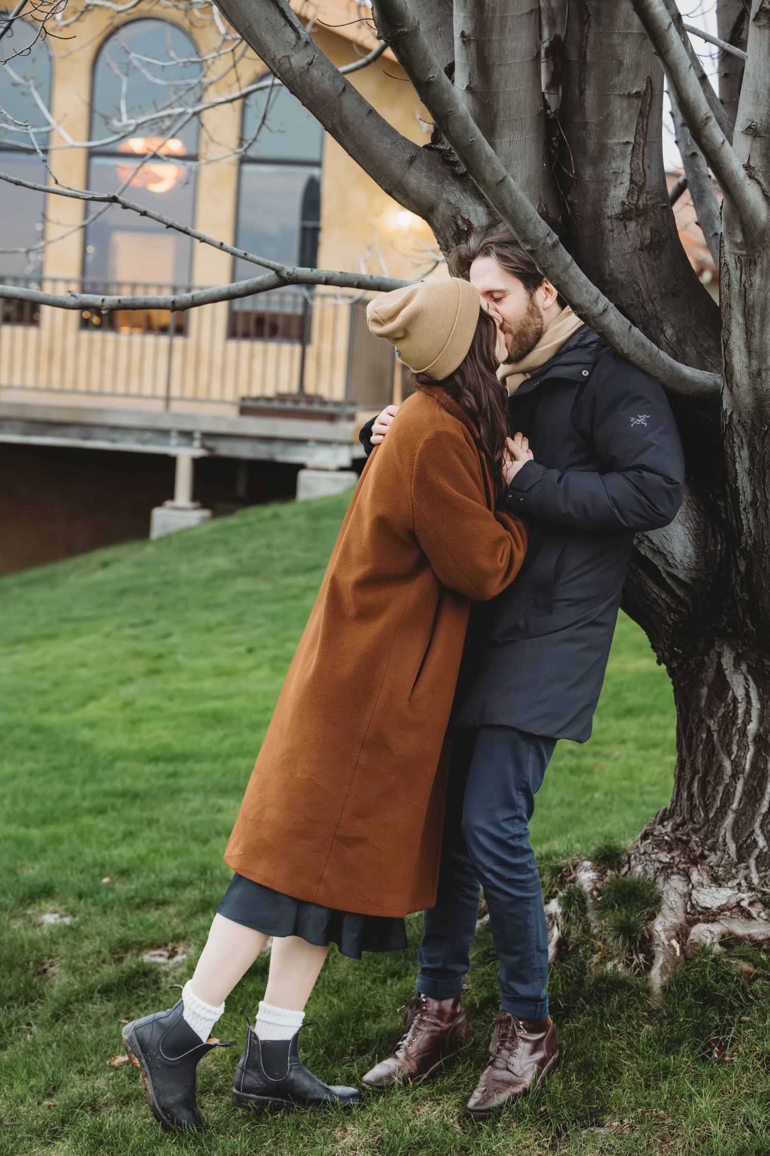 A man leaning against a tree with a winery in the background as his new fiancee gives him a kiss.