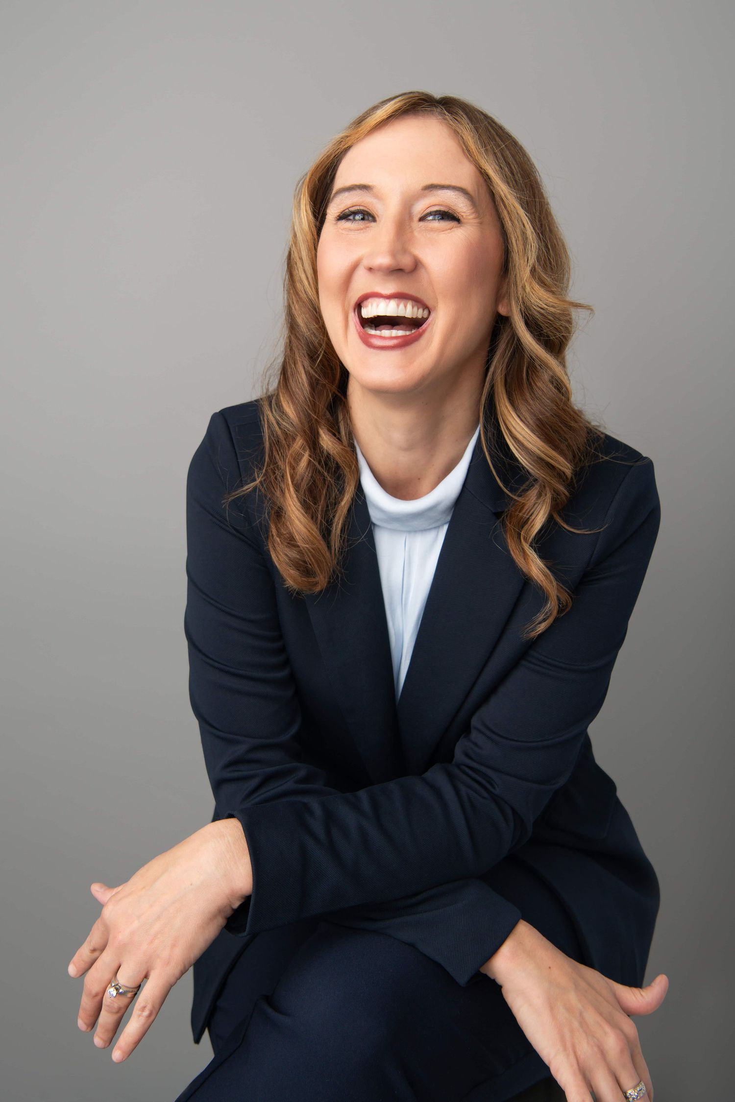 Studio headshot of a professional woman laughing naturally while seated against a neutral gray backdrop in a tailored blazer.