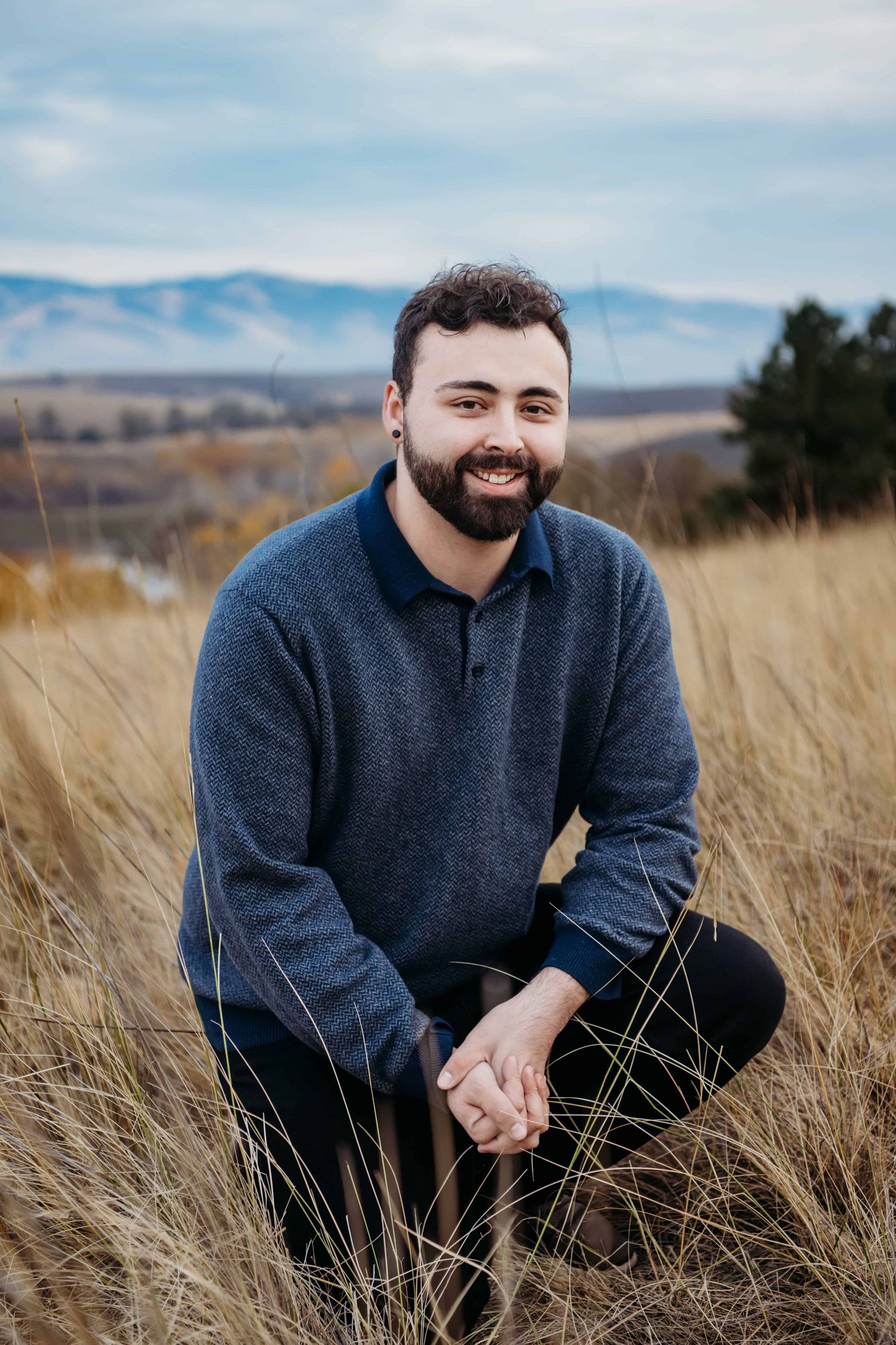 A portrait of a man kneeling in tall neutral colored grasses with rolling hills in the background.