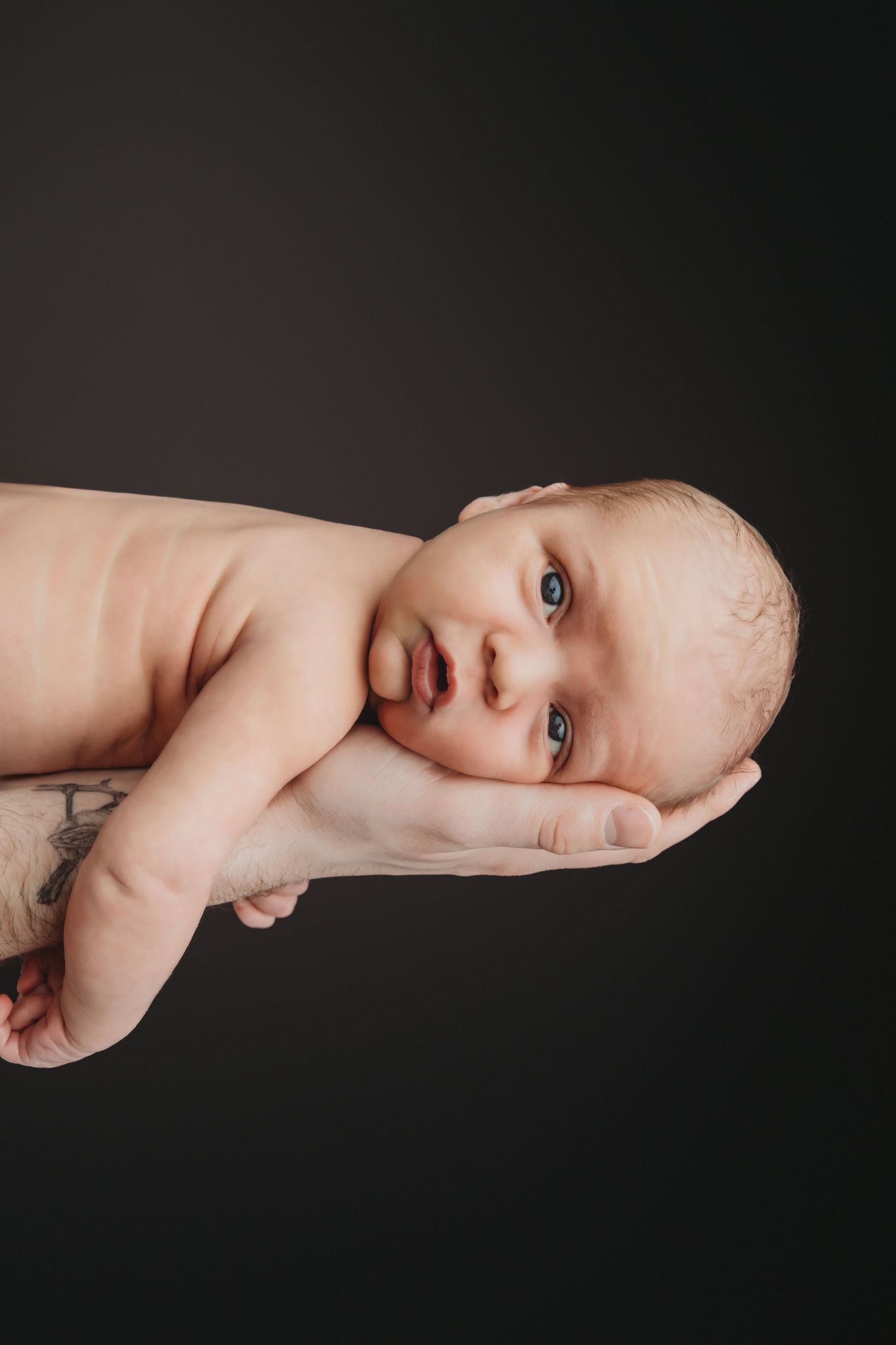 A newborn laying in his dad's hand, looking at the camera on a black backdrop.