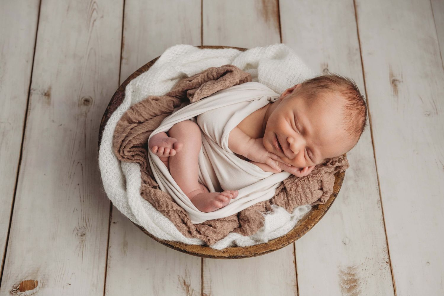 A baby boy wrapped in white sleeping in a brown wooden bowl on white rustic flooring.