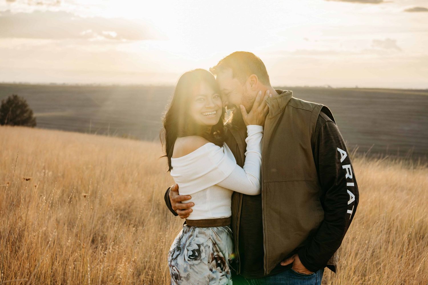 A man cuddling his wife while she looks at the camera and smiles during golden hour.