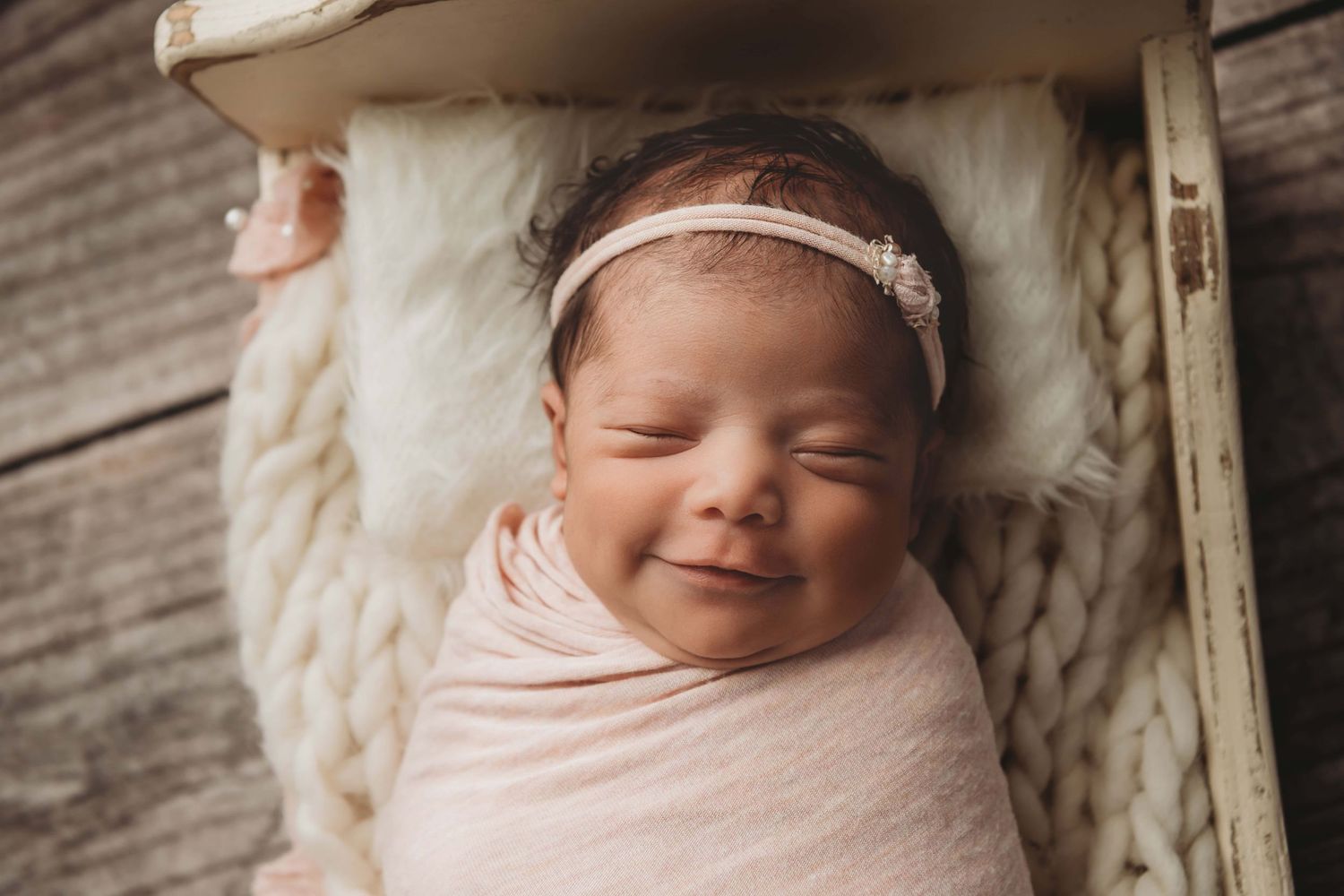 A newborn smiling with her eyes closed in a bed with cream and soft pinks.