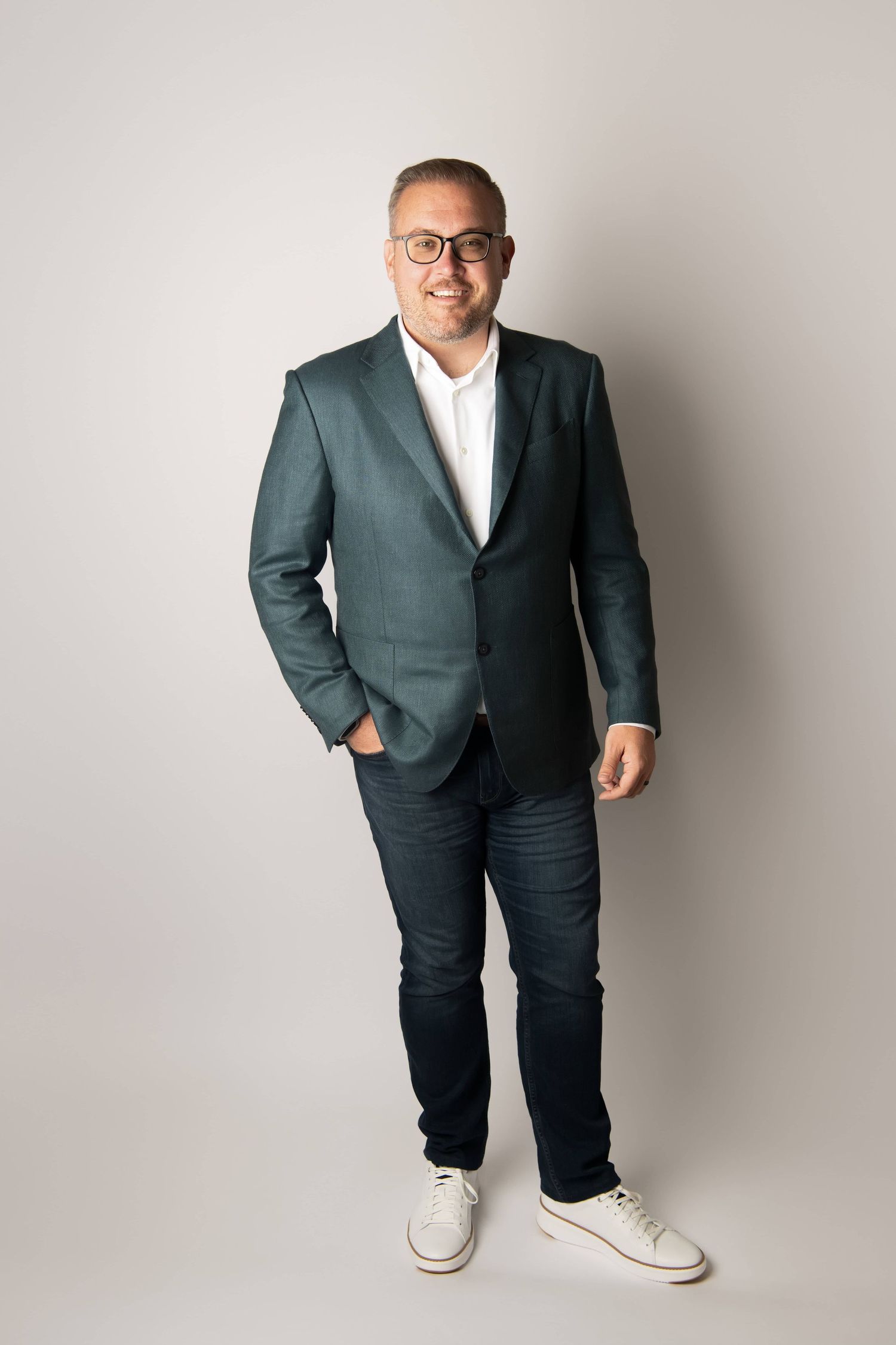 A full body portrait of a man wearing a suit jacket on a white backdrop.