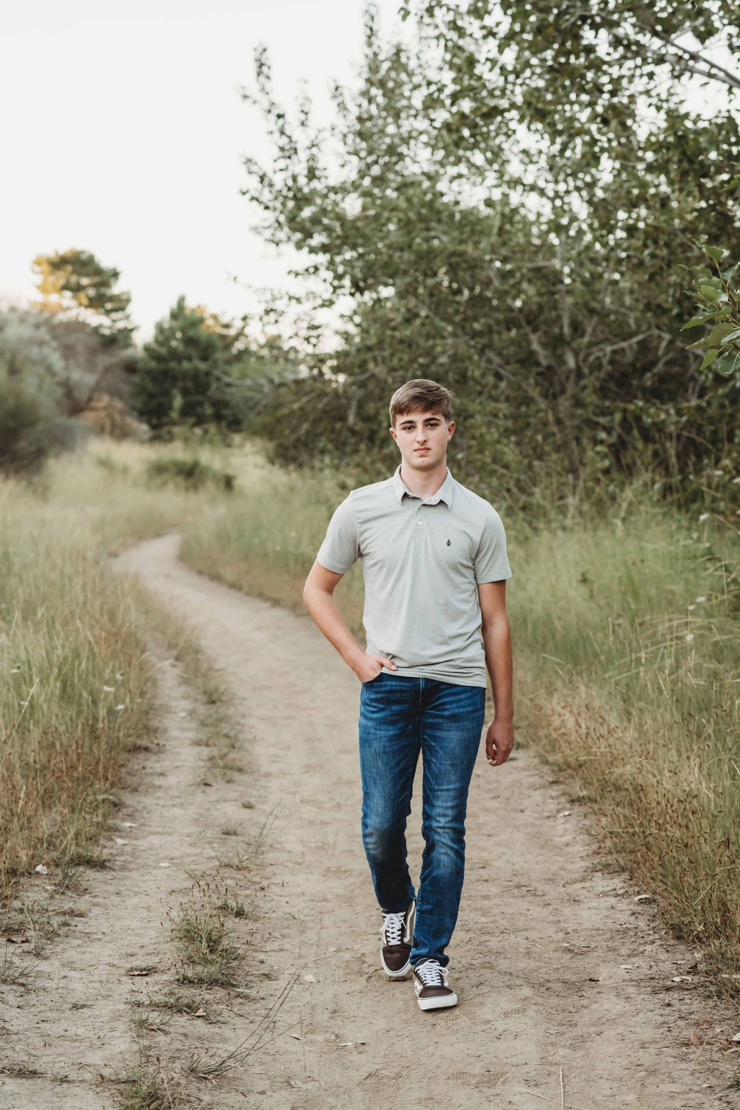 A young man walking down a dirt path with his hand in his pocket, looking at the camera with a serious expression.