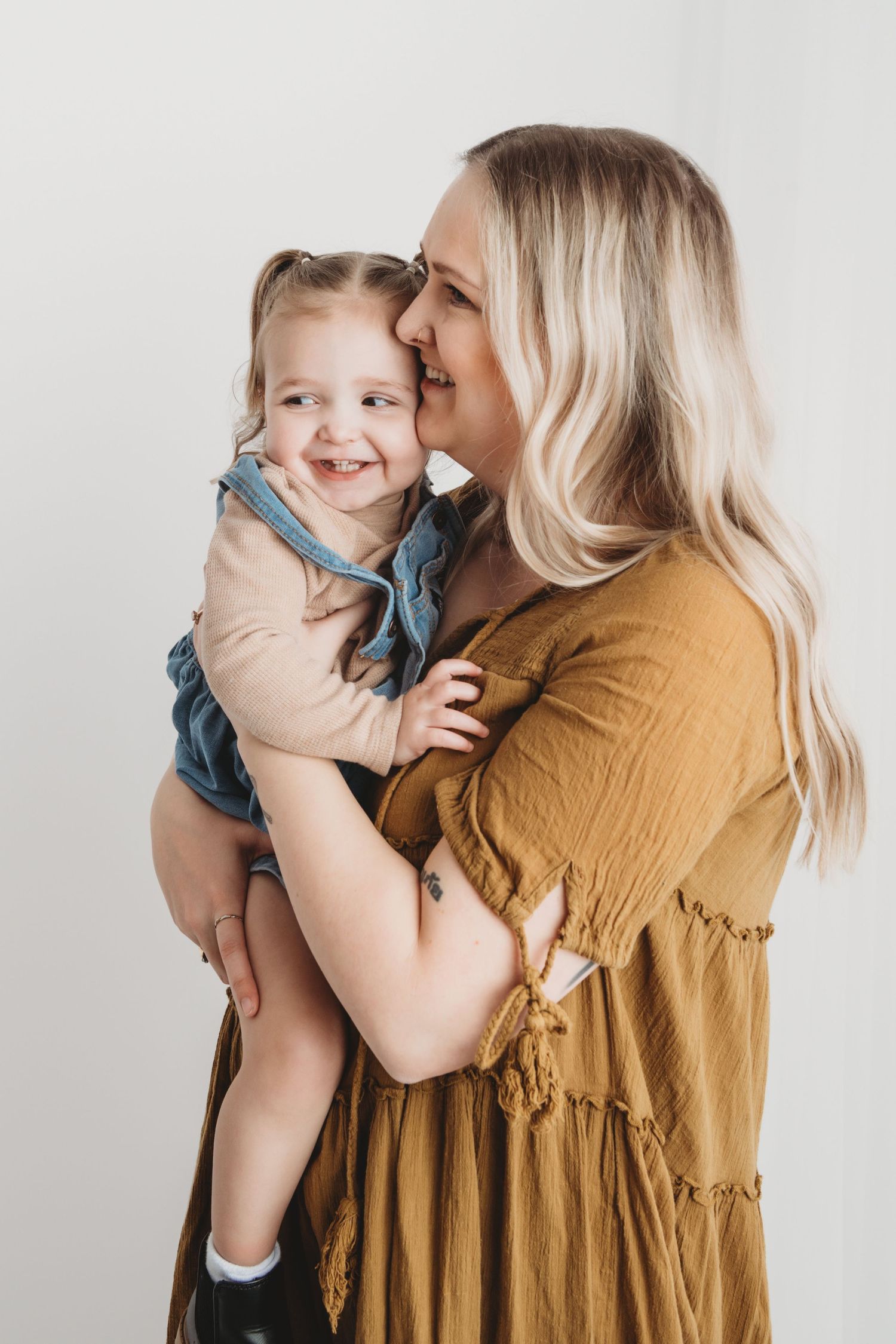 A mom and her toddler girl hugging and laughing on a white backdrop.