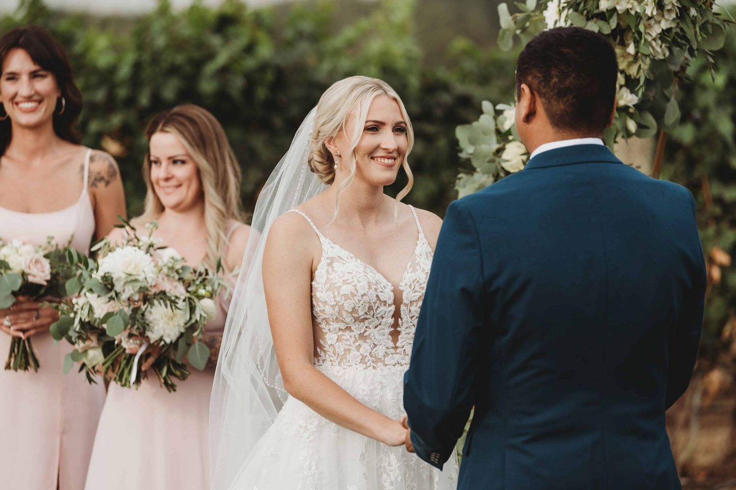 A bride looking at her groom and smiling during their wedding ceremony, with bridesmaids smiling in the background.