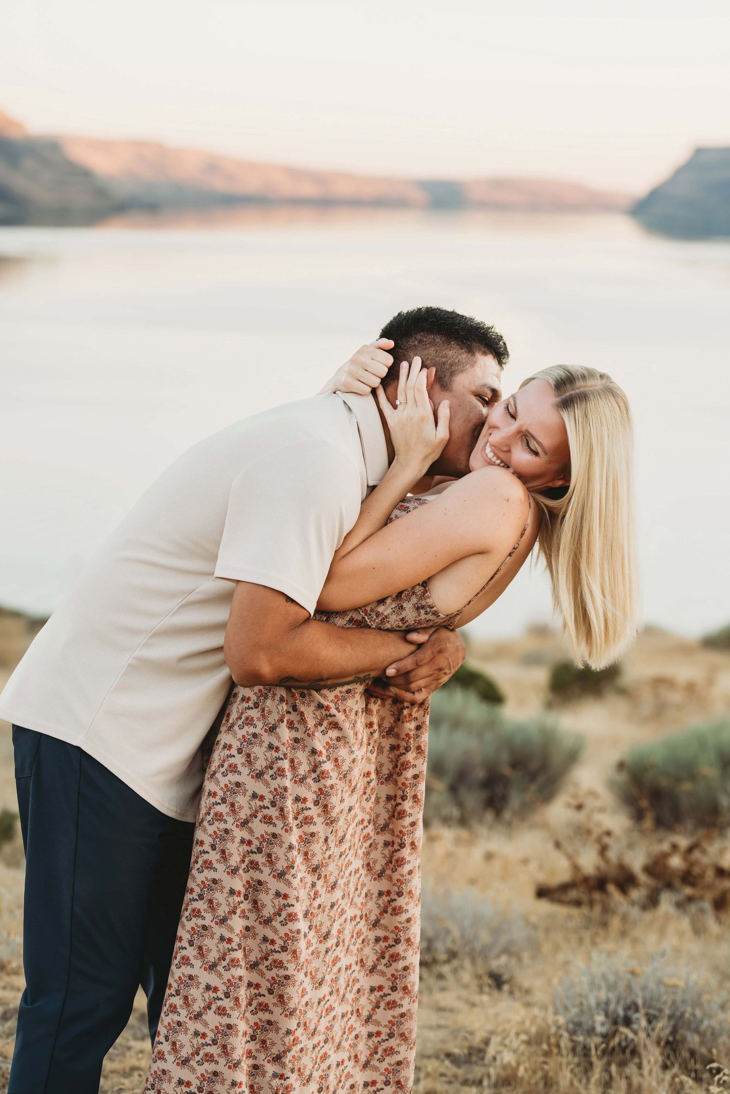 A man playfully kissing his fiancé's neck with a lake in the background.