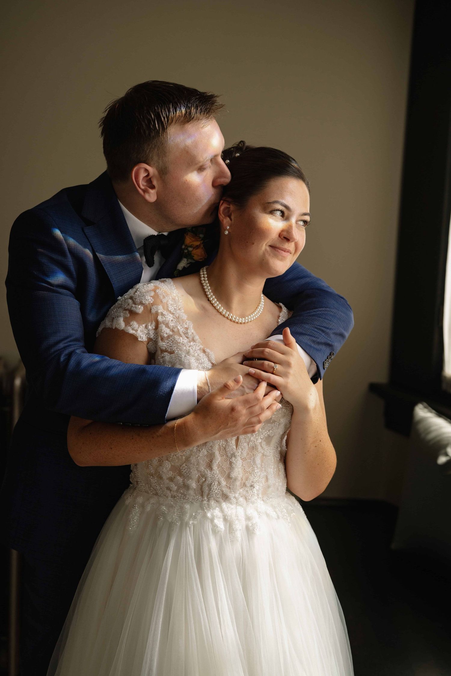 A window lit image of a groom kissing his bride on her head as she holds his hands.